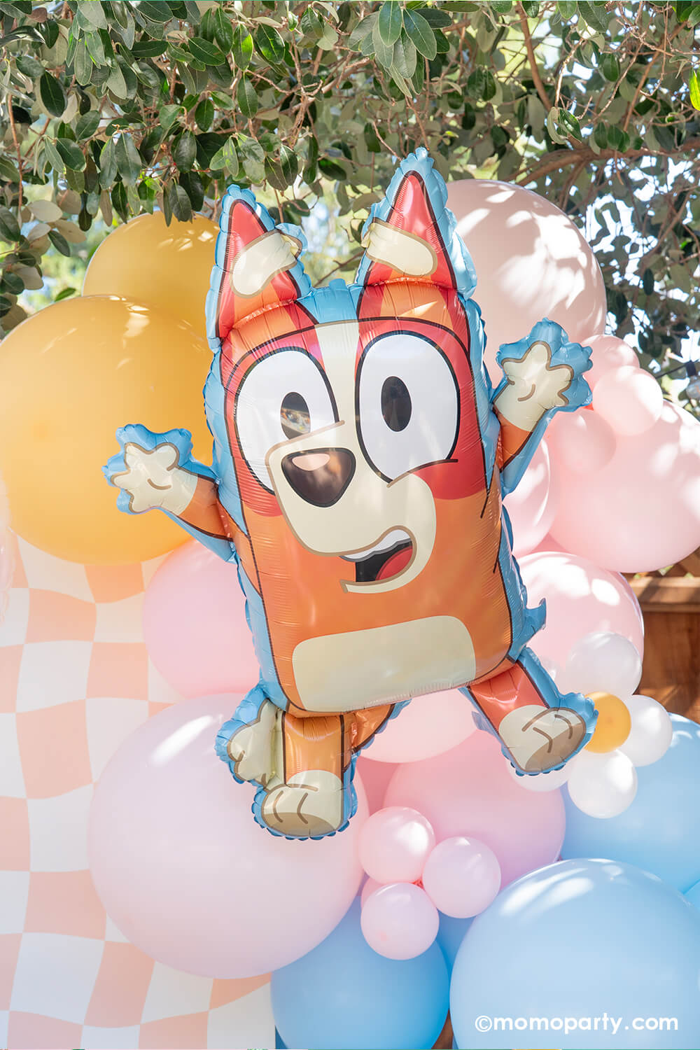 Momo Party's 23" x 32" Bluey Bingo shaped foil balloon among a Bluey themed balloon garland in pastel peach, pink and blue adorned with daisy balloons with a wavy peach checkered party backdrop board at a kid's birthday party setting in a backyard.