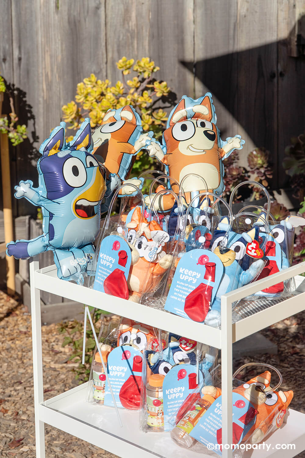 A kid's Bluey themed birthday party favor white cart filled with colorful Bluey themed favors including Bluey and Bingo mini stuffies, red balloons, and Bluey and Bingo mini shaped foil balloons from Momo Party with flowers in the background.