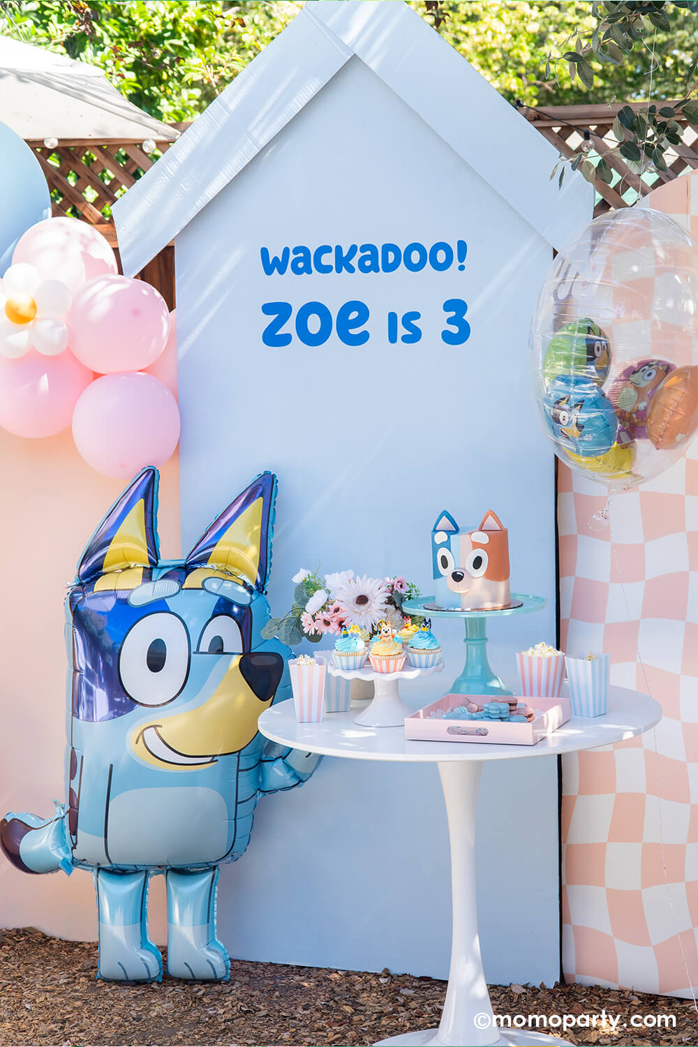 An adorable kid's Bluey themed birthday party setup with a 'Wackadoo! Zoe is 3' on a light blue backdrop board along with two pastel peach boards adorned with pastel colored balloon garlands. In the front, there's an cute jumbo 36" Bluey AirWalkder foil balloon from Momo Party standing next to a dessert table with a Bluey themed birthday cake, cupcakes, sweets, desserts in matching pastel colors of blue and peach.