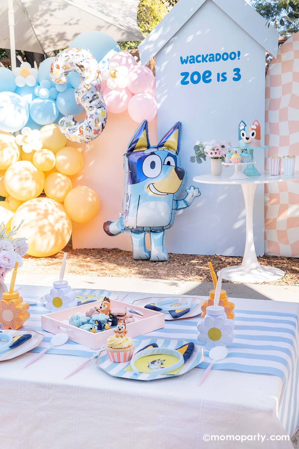 An adorable party decoration set of by Momo Party for a kid's Bluey themed birthday party featuring pastel blue and peach backdrop boards with 'Wackadoo! Zoe is 3' adorned with matching colored balloon garland and a Bluey number three balloon. In the front, there's the jumbo 36" Bluey AirWalkder foil balloon from Momo Party standing next to a dessert table with a Bluey themed sweets. In the front set a kids picnic table with Bluey themed tableware including plates, napkins, party cups from Momo Party.