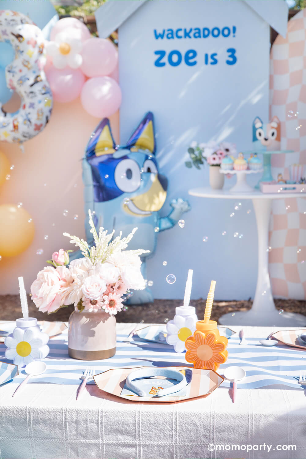 An adorable party decoration set of by Momo Party for a kid's Bluey themed birthday party featuring pastel blue and peach backdrop boards with 'Wackadoo! Zoe is 3' adorned with matching colored balloon garland and a Bluey number three balloon. In the front, there's the jumbo 36" Bluey AirWalkder foil balloon from Momo Party standing next to a dessert table with a Bluey themed sweets. In the front set a kids picnic table with Bluey themed tableware including plates, napkins, party cups from Momo Party.