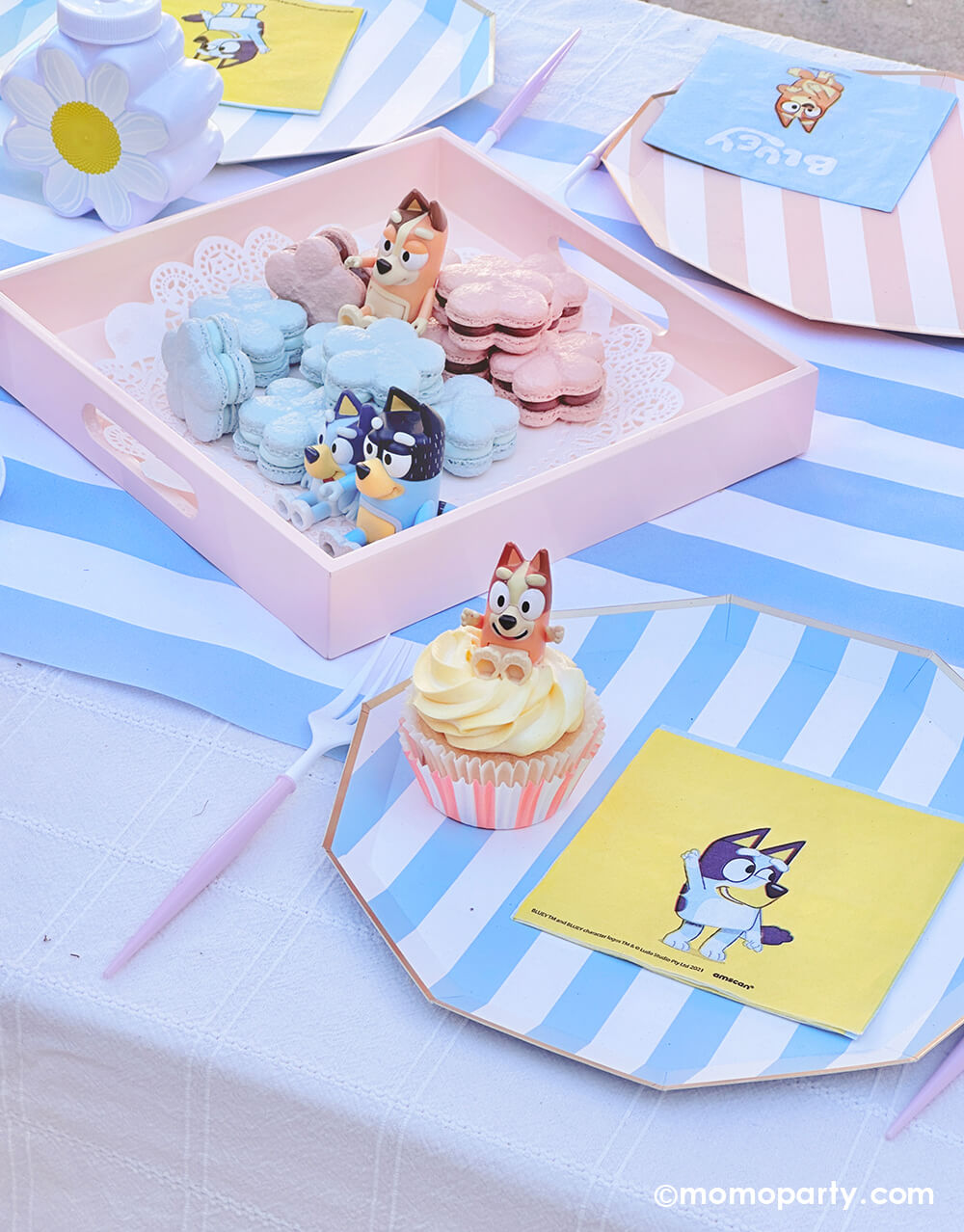 An adorable kid's picnic party table setting with Bluey themed various tablewares from Momo Party including summer blue striped dinner plates, light blue striped table runner, daisy shaped tumbler cups in peach and white, Bluey small napkins, Bluey decorative headbands. Along with a Bluey-inspired daisy shaped macarons, floral bouquet decorations, it's a perfect table setting idea for a modern Bluey themed birthday celebration.