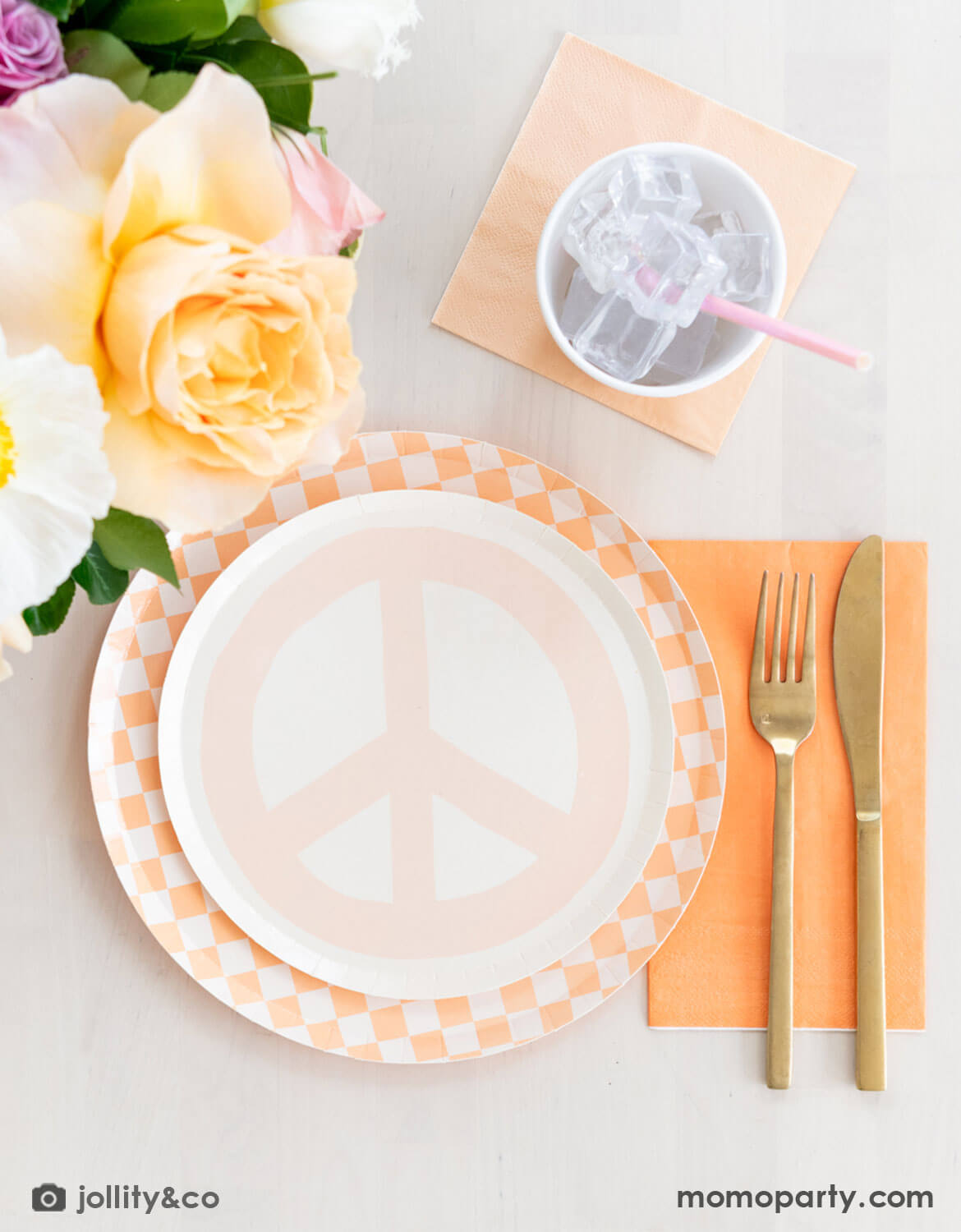 An elegant table setting featuring a pastel peach peace symbol plate with a peach checkered dinner plate, orange and peach napkins, flowers, and cutlery from Momo Party on a light surface.
