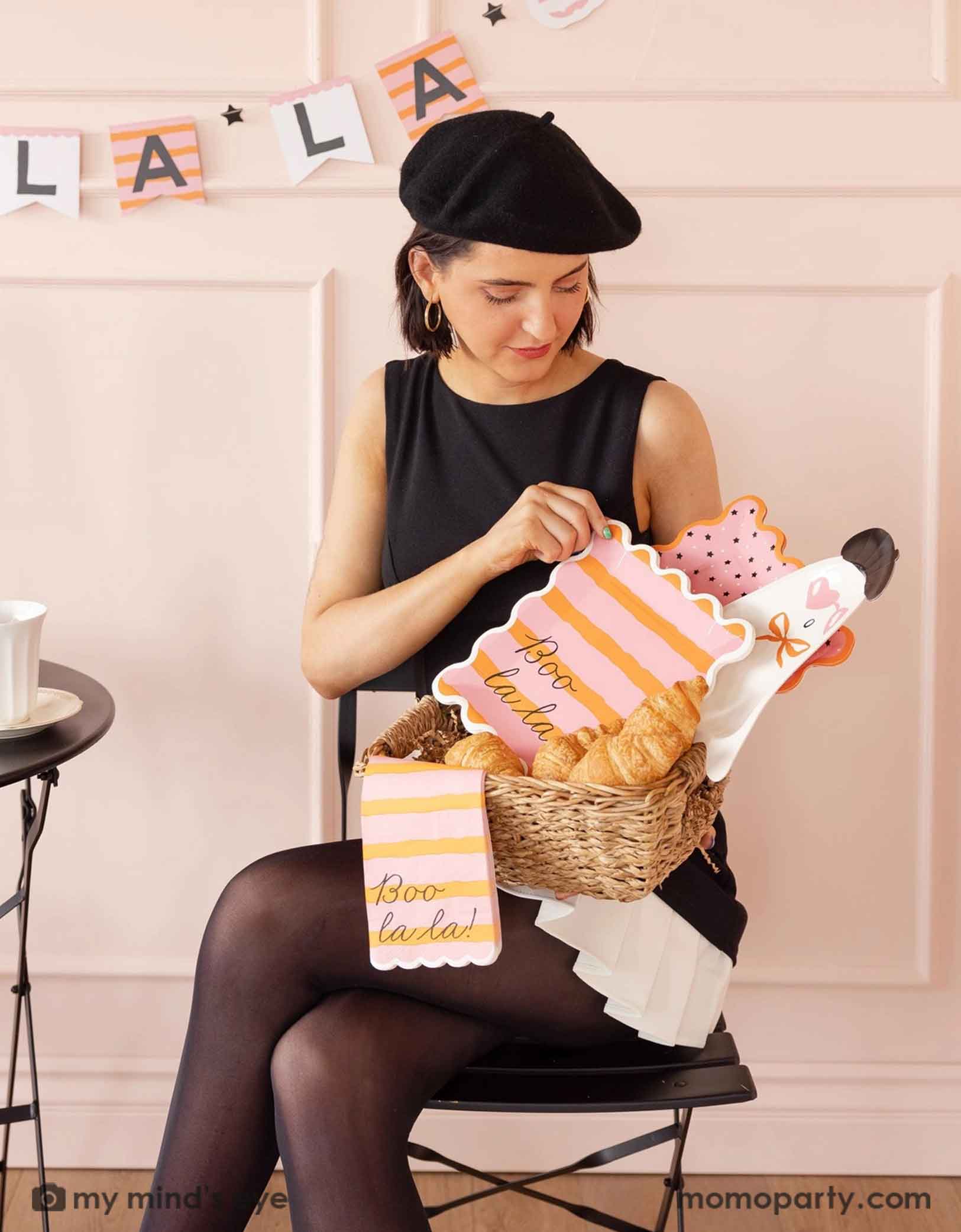 A lady in a black dress and beret holding a basket with Momo Party's Boo La La french inspired Boo La La party tableware collection featuring the ghost-shaped plate, the orange and pink striped plate and napkins the at a Halloween-themed party decorated with french-inspired striped Boo La La party banner on the wall.