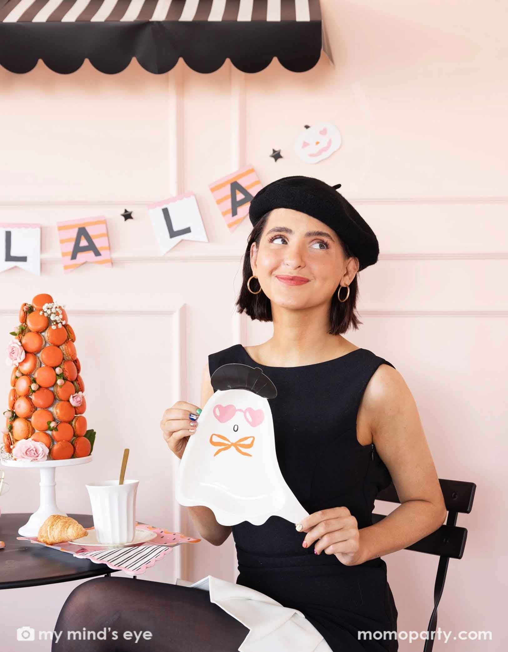 A lady in black dress and beret holding Momo Party's Boo La La french inspired ghost-shaped plate at a Halloween-themed party decorated with french-inspired party supplies including striped Boo La La party banner, striped napkins, and decorations, , along with a macaron tower on the table.
