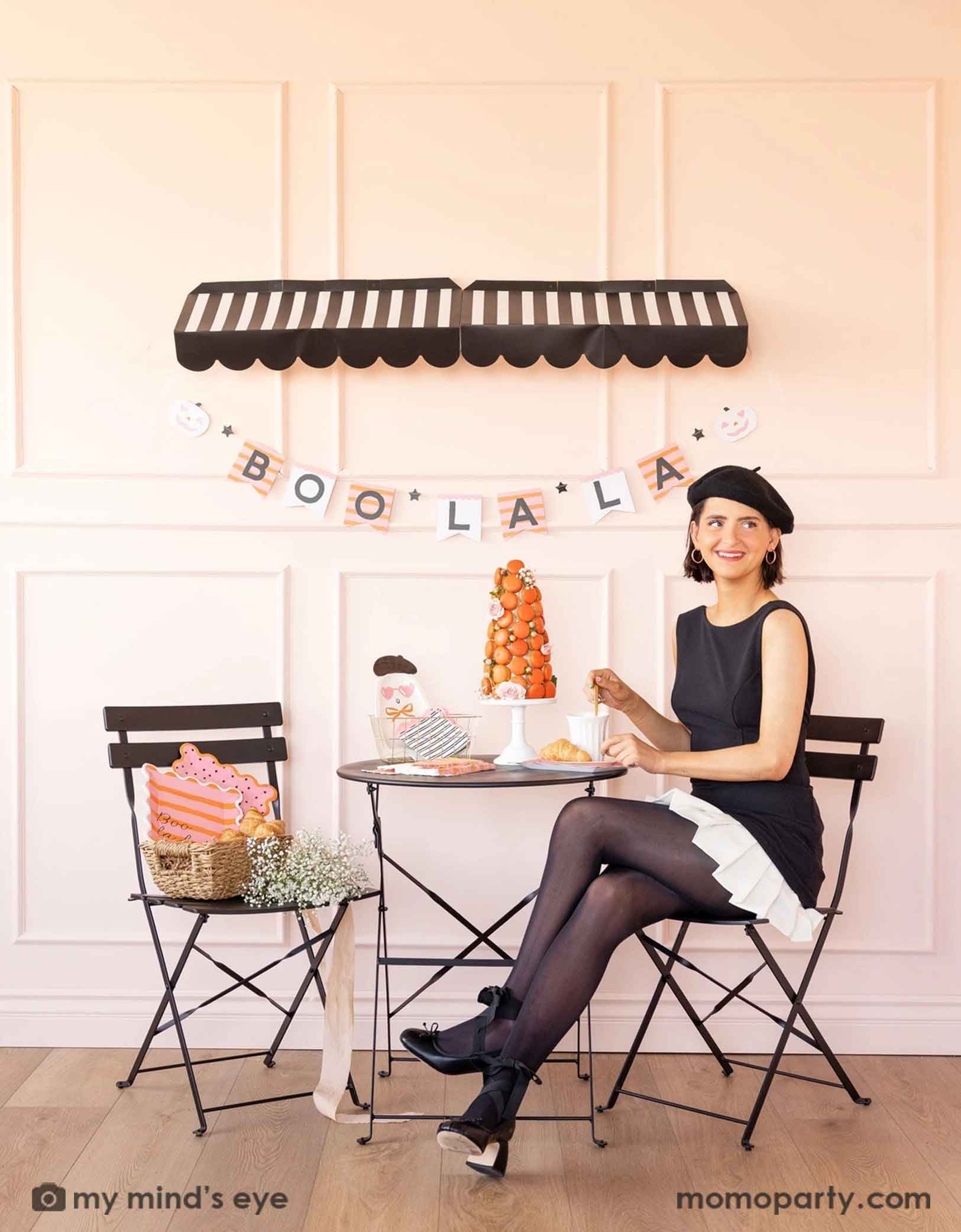 A lady in black dress and beret sitting in a French inspired pink Halloween set up featuring Momo Party's Boo La La party collection which includes ghost-shaped plates, black striped with pink edge napkins, orange and pink striped plates and in a basket with croissants and flowers and a macaron tower on the table. In the background there's a black striped awning and underneath hung the Boo La La striped party banner. A chic idea for a not-so-spooky ghost themed Halloween celebration for girls gathering.