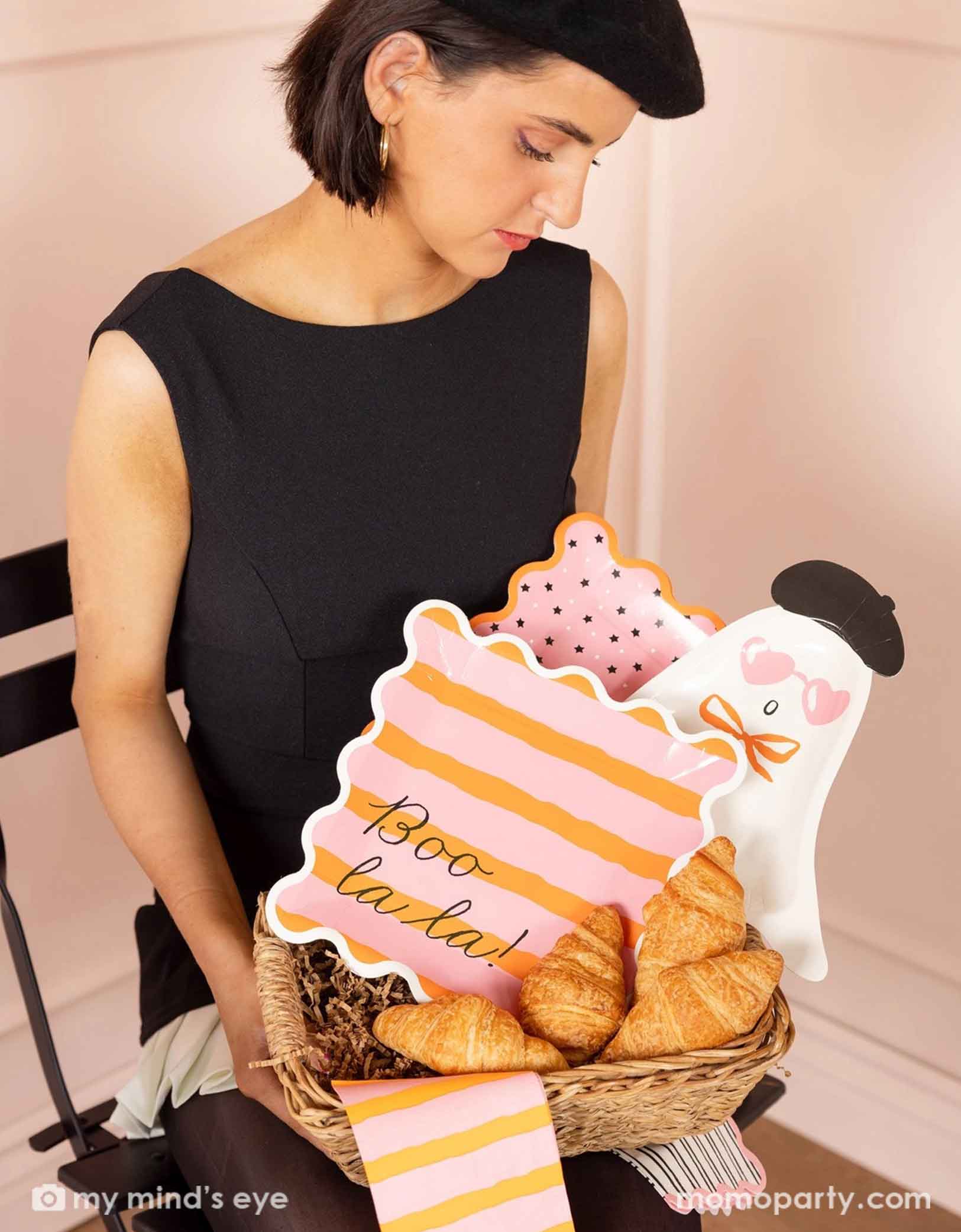 A lady in a black dress and a beret hat holding a french inspired basket filled with Momo Party's Boo La La party tableware collection including the ghost shaped plates, the orange and pink striped dinner plates & napkins, and the pink and black star plates, with croissants around the tablewares.