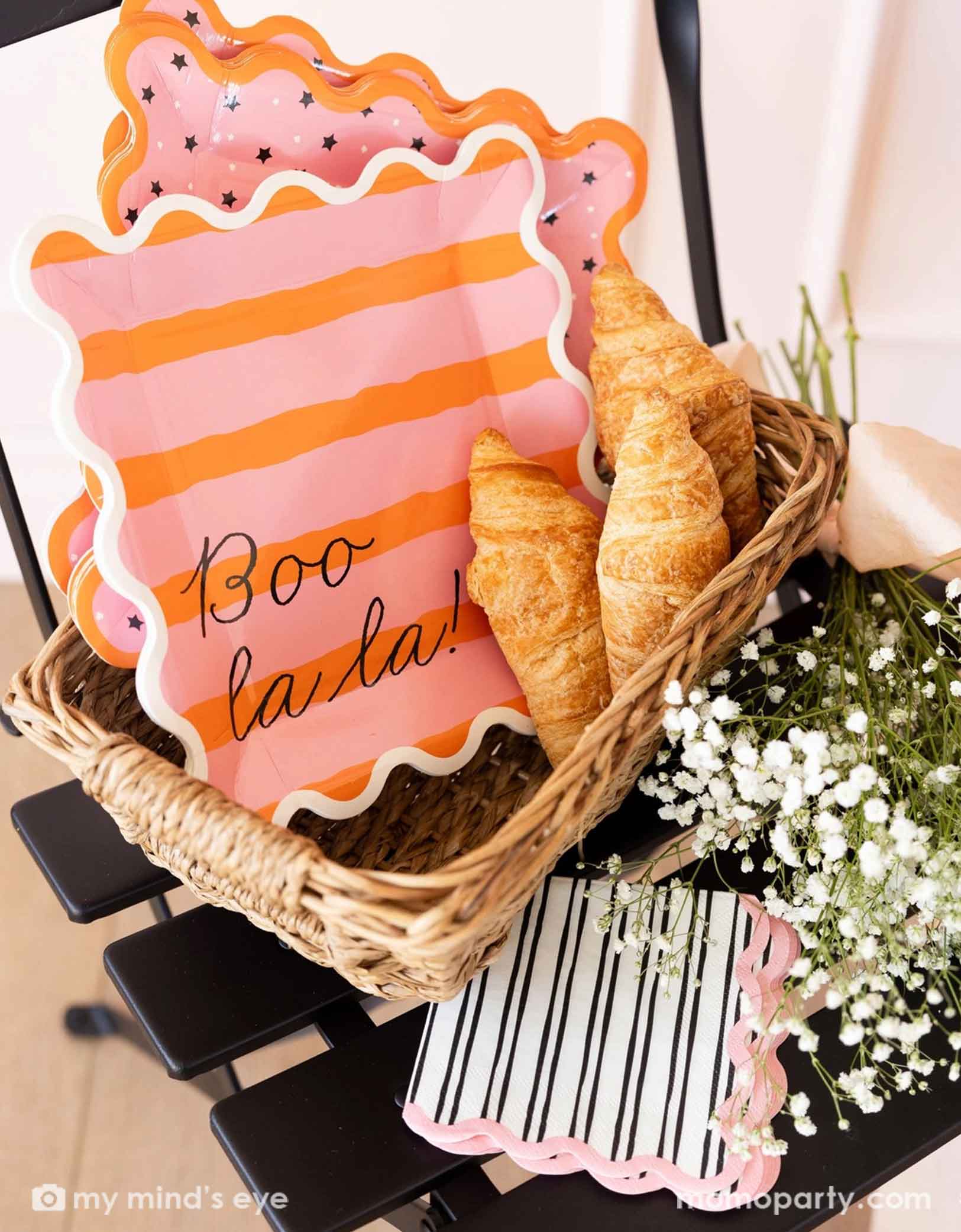 A french inspired basket filled with Momo Party's Boo La La party tableware collection including the orange and pink striped dinner plates, the pink and black star plates, black and pink striped napkins and with pastries and flowers around it.