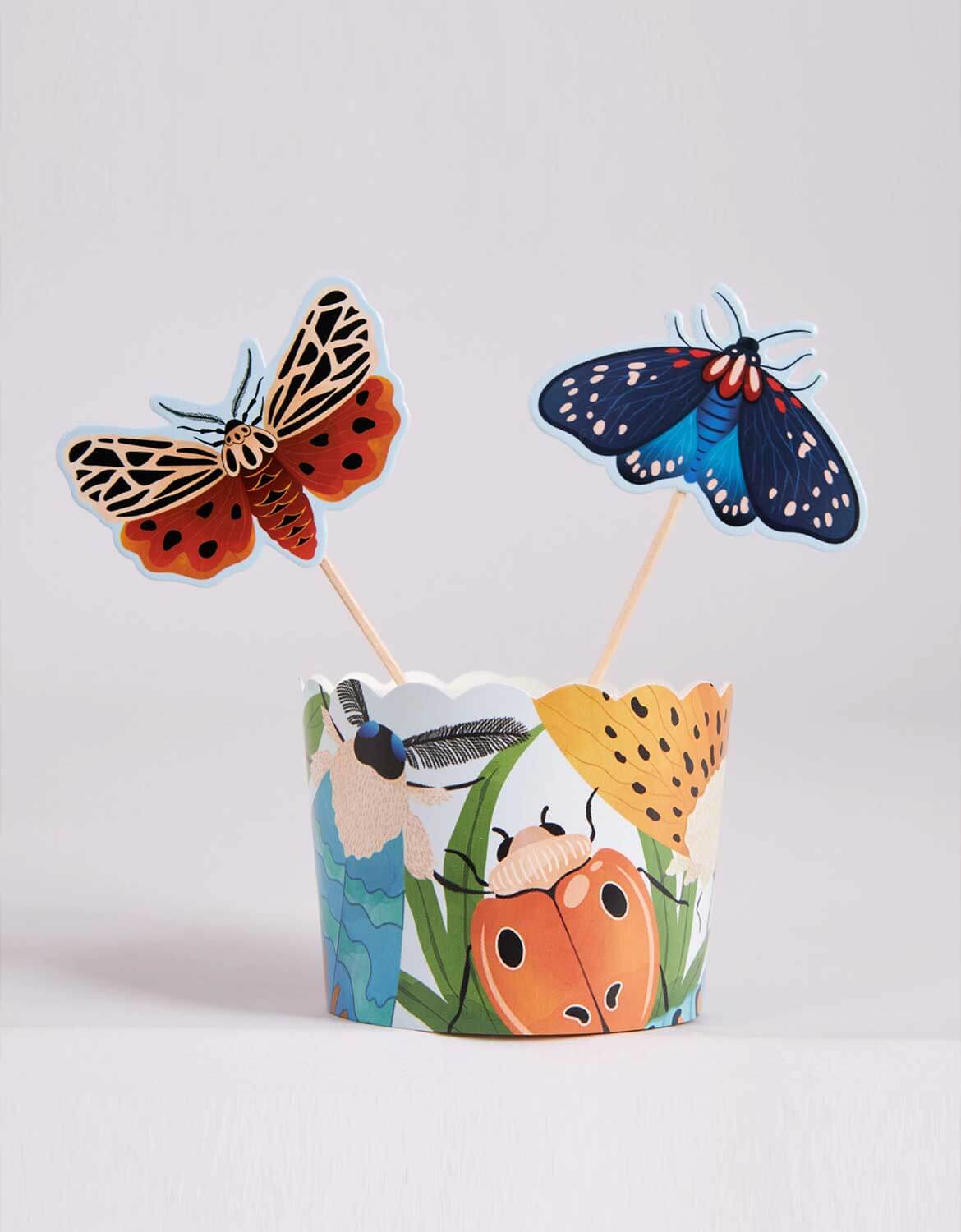 Two colorful butterfly-themed cupcake toppers with a decorative insect themed cupcake wrapper from Momo Party against a light gray background.