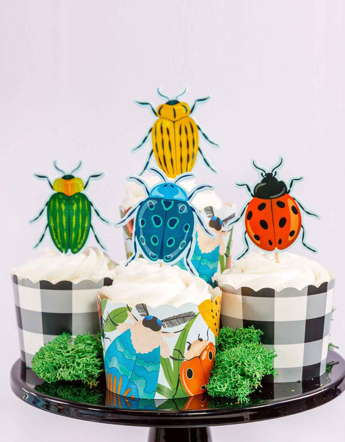 Four cupcakes with insect-themed cake toppers and wrappers from Momo Party on a black cupcake stand against a white background.