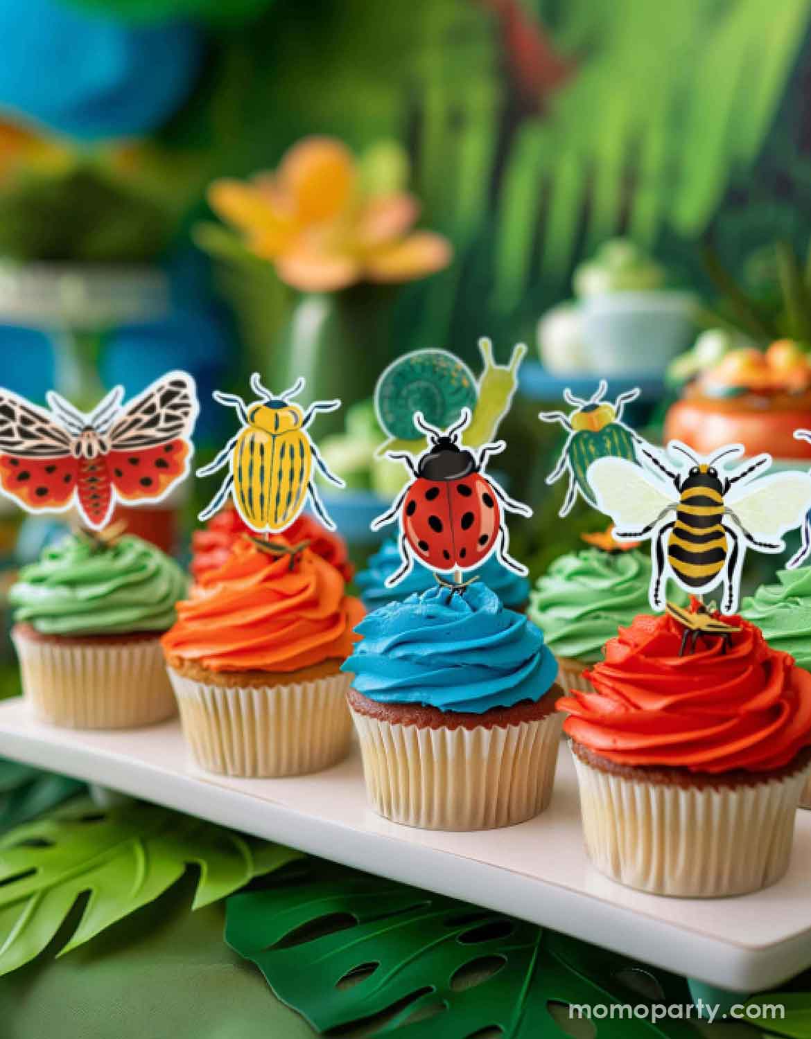 A set of festive cupcakes with colorful frosting and insect-themed toppers from Momo Party on a decorative natural themed background with palm leaves and tropical flowers.