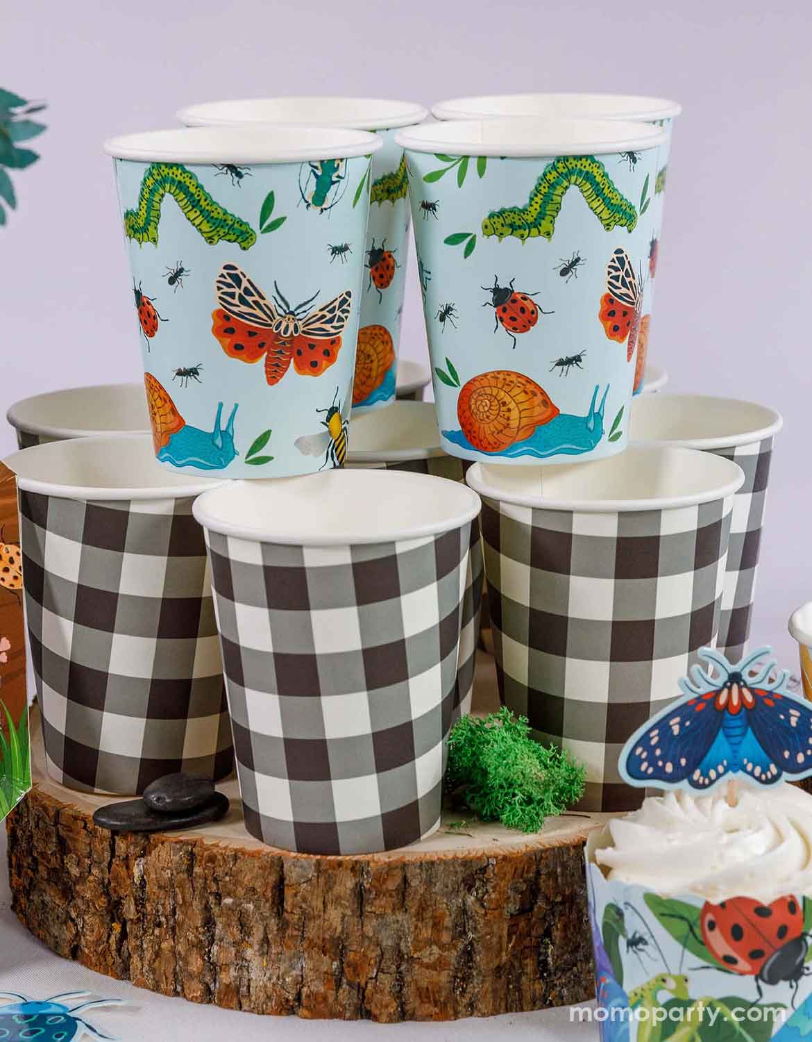 A set of checkered paper cups and light blue party cups with insect-themed designs from Momo Party on a wooden stand. In the front there's a cupcake topped with an insect topper and wrapper.