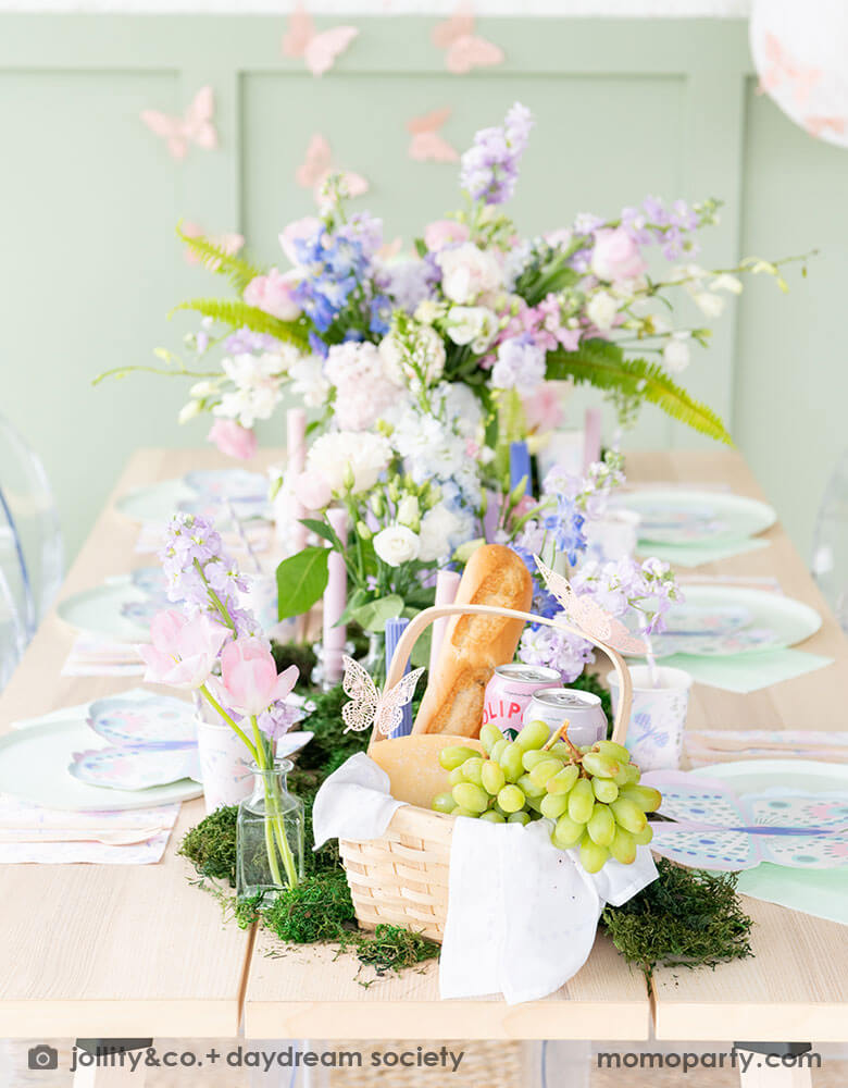 A beautiful butterfly themed party table featuring Momo Party's Flutter butterfly shaped plates paired with pastel mint dinner plates and napkins, next to it is butterfly themed party cups and napkins. In the middle there's moss grass textured table runner with pretty spring flower arrangement and a picnic basket of fruits and breads. On the wall there are butterfly shaped paper decorations attached on the wall, making this a perfect inspo for kid's butterfly fairy themed birthday or a spring celebration.