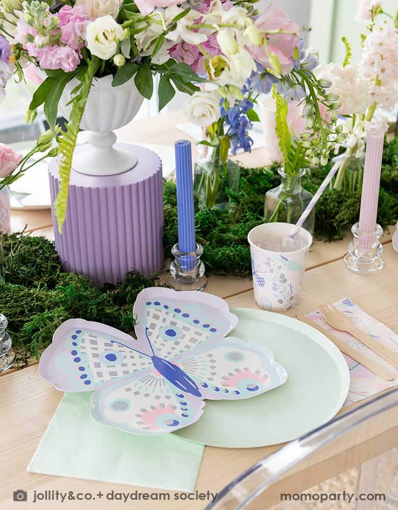 A beautiful butterfly themed party table featuring Momo Party's Flutter butterfly shaped plates paired with pastel mint dinner plates and napkins, next to it is butterfly themed party cups and napkins. In the middle there's moss grass textured table runner with pretty spring flower arrangement, making this a perfect inspo for kid's butterfly fairy themed birthday or a gorgeous spring Easter celebration.
