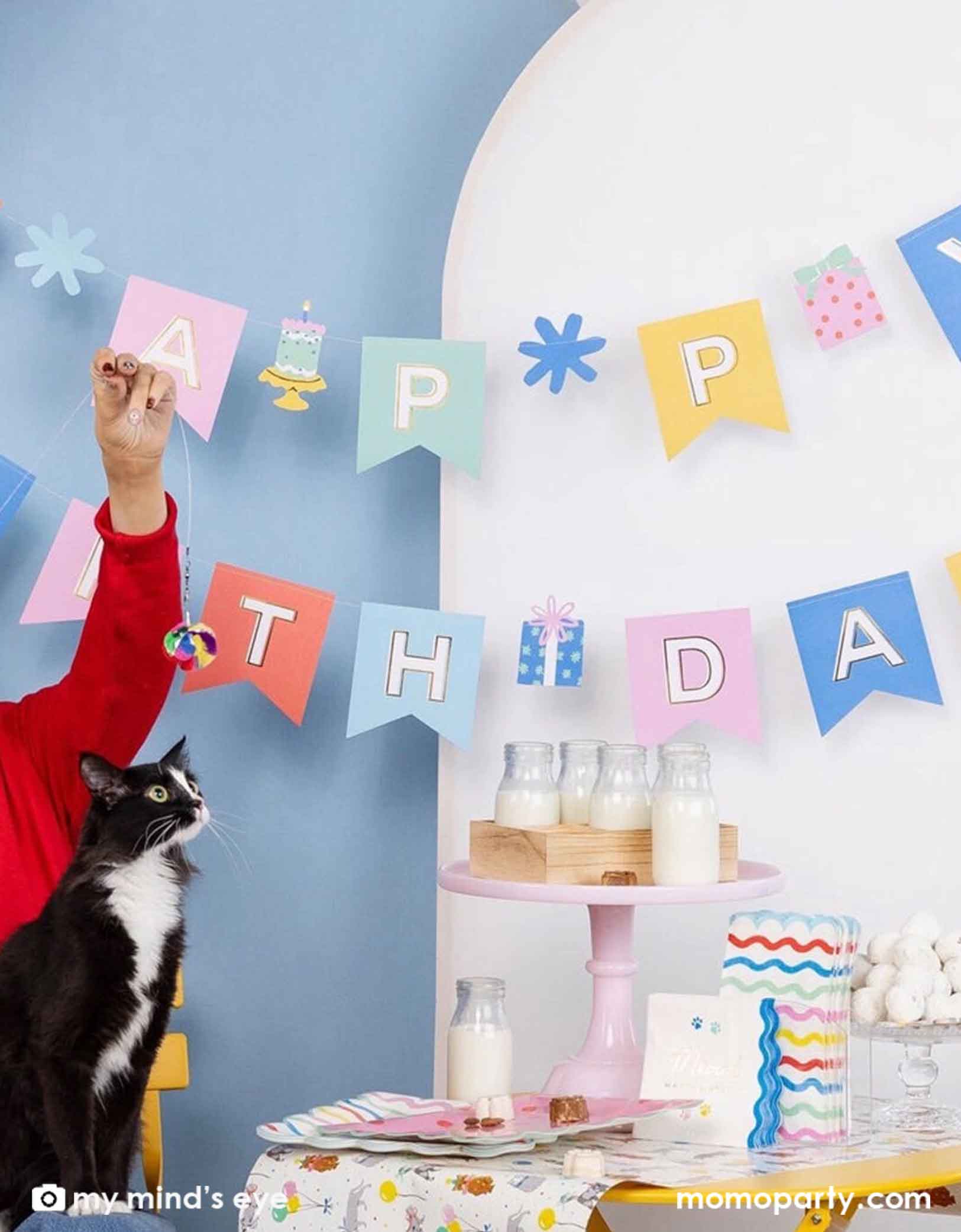 A cat themed birthday party setup with Momo Party's 5ft Cat Happy Birthday party banner, milk bottles, and a table of cat themed party supplies and a black and white cat looking at the camera.
