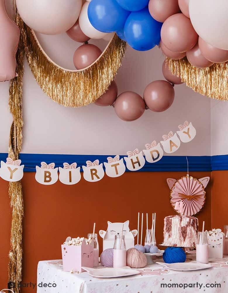 A kid's cat themed birthday party decoration set up features Momo Party's cat themed party supplies by Party Deco including 18" kitty cat shaped foil balloon in cream, along with some latex balloon mix in pink, blush and royal blue. On the wall there are pink kitty cat shaped party fans along with a gold tinsel garland hung on. With a pink number 4 foil balloon and a cat head shaped happy birthday banner, it makes an adorable inspo a cat or pet themed party.
