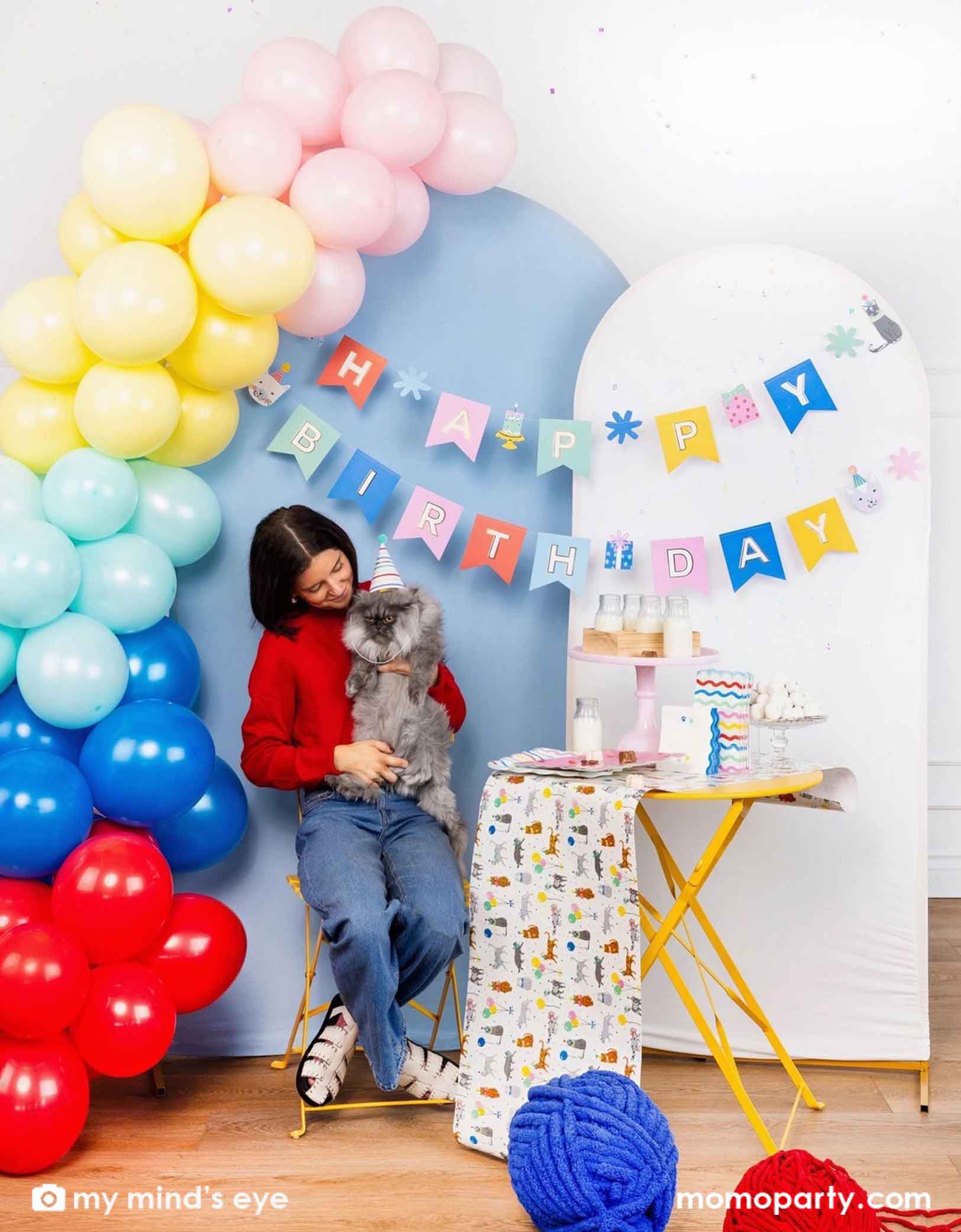 A woman holding a flurry cat that hears a birthday hat in front of a colorful balloon arch with Momo Party's Cat 'Happy Birthday' party banner hung on the blue and white party backdrop boards. Next to it is a set of yellow table and chair filled with cat themed party supplies from Momo Party including plates, napkins, table runner, and party cups. Along with the jumbo yarns in blue and red on the ground, this sets a great idea for cat themed parties.