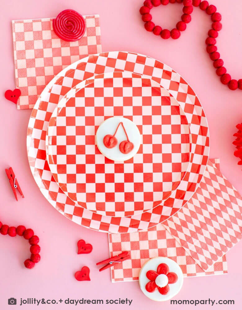 A pink and red party table featuring Momo Party's check it red checkered party collection including red checkered patterned dinner and side plates, large and small napkins by Jollity Co. With sugar cookies with red flower and cherry designs and some party favors goods and felt garland around the tableware, it makes it a modern and chic playful party table.