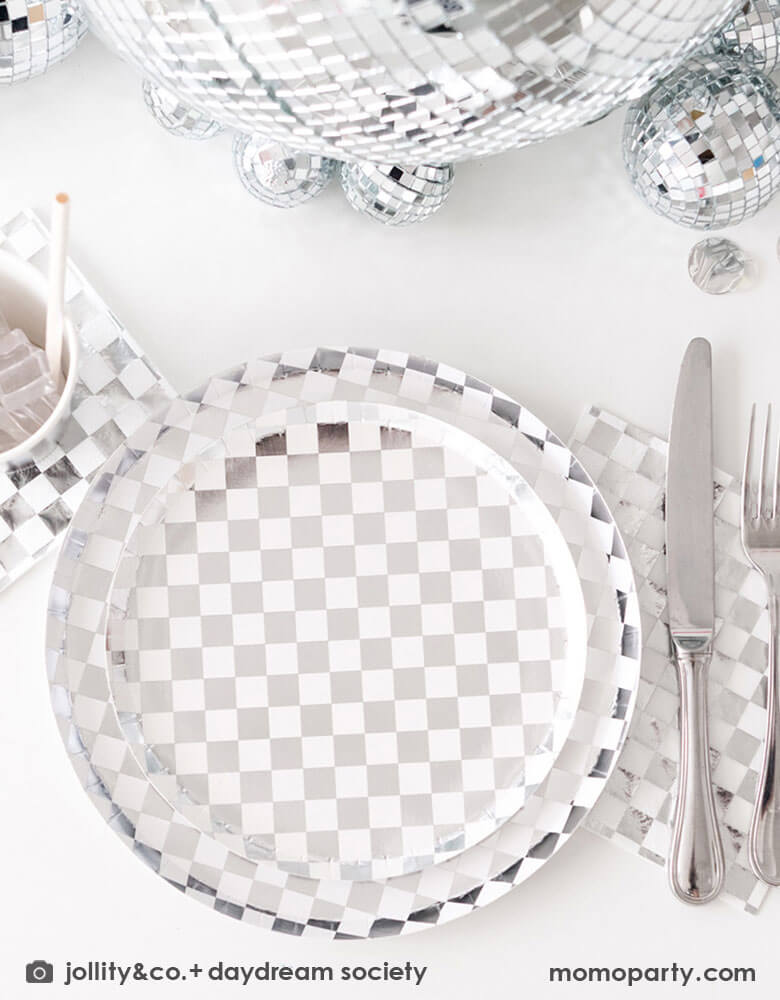 A glamorous and dazzling party table featuring Momo Party's check it! silver checkered tableware collection by Jollity & Co. including 10" dinner plates, 8" dessert plates, 6.5" large napkins, solid silver party cups and silver cutlery. In the middle of the table are a bunch of disco balls in different sizes with silver confetti scattered, what a festive party table! A perfect inspo for New Year's Eve celebration or a disco themed party!