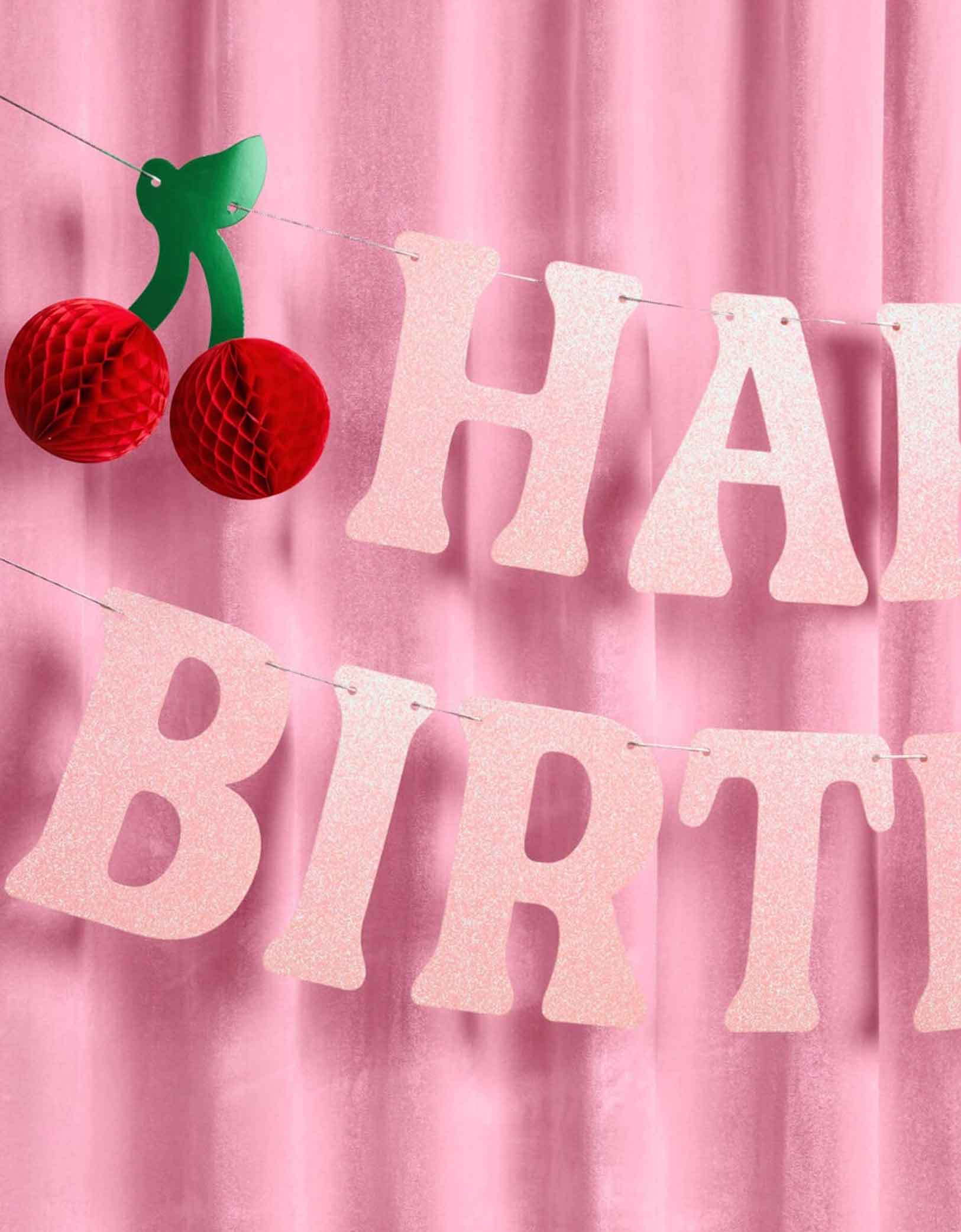 A close up of Momo Party's 4.5' cherry happy birthday banner hung against a pink curtain.
