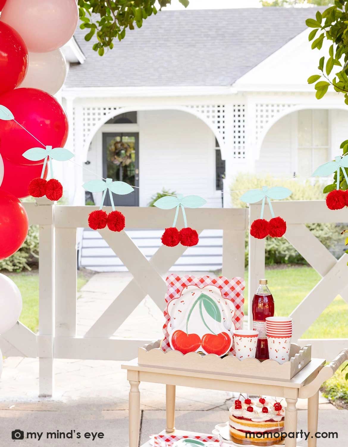 An outdoor party setting next to a red and pink balloon garland with cherry-themed decor and party tableware from Momo Party including cherry themed plates, cups, a bottle of cherry juice and a cherry cake on a wooden serving cart. Above it hung a pom pom cherry felt party banner - a simply yet adorable idea for am outdoor cherry themed party in summer.