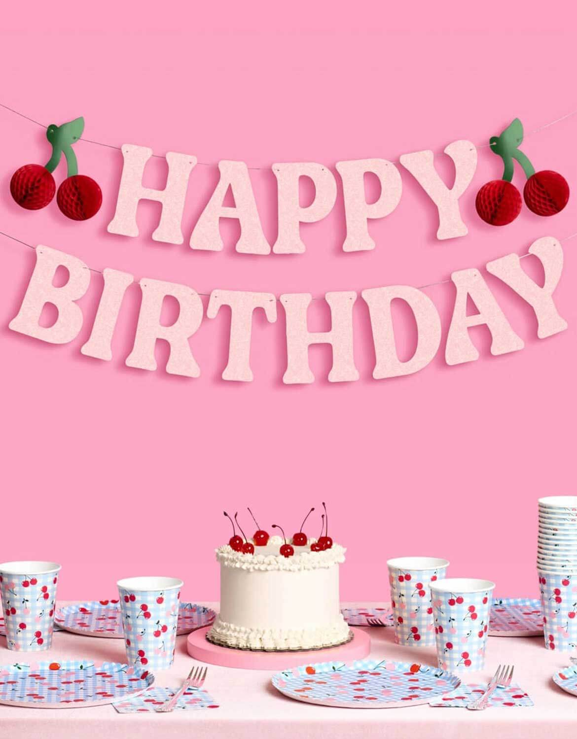 Momo Party's 4.5' cherry happy birthday banner hung above a table full of cherry with blue gingham themed party tablewares, against a pink background.