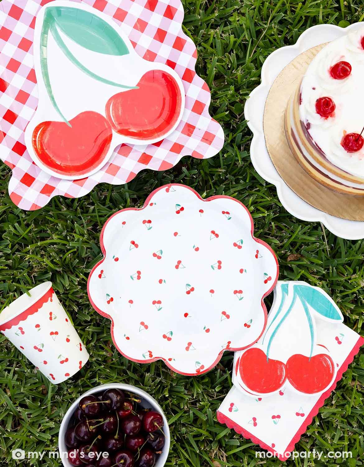 Momo Party's cherry-themed party plates, party cups and napkins by My Mind's Eye on grass in a picnic setting with a cake, and a small bowl of cherries around.