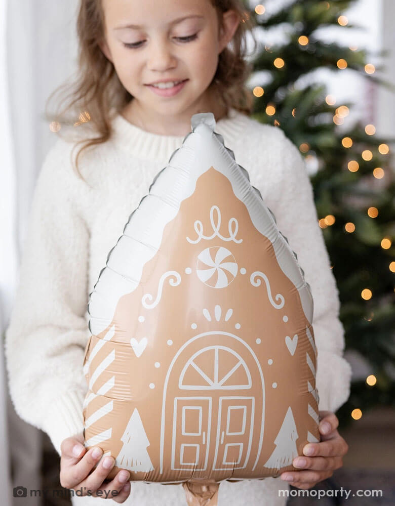 A girl in her cream white sweater holding Momo Party's 13.5" x 20" gingerbread house shaped foil balloon standing next to a big window. Behind her is a big Christmas tree lit up with small twinkling lights,