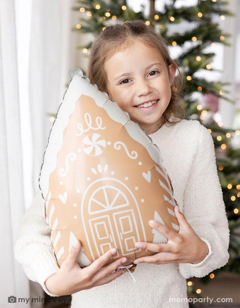 A girl in her cream white sweater holding Momo Party's 13.5" x 20" gingerbread house shaped foil balloon standing next to a big window. Behind her is a big Christmas tree lit up with small twinkling lights,