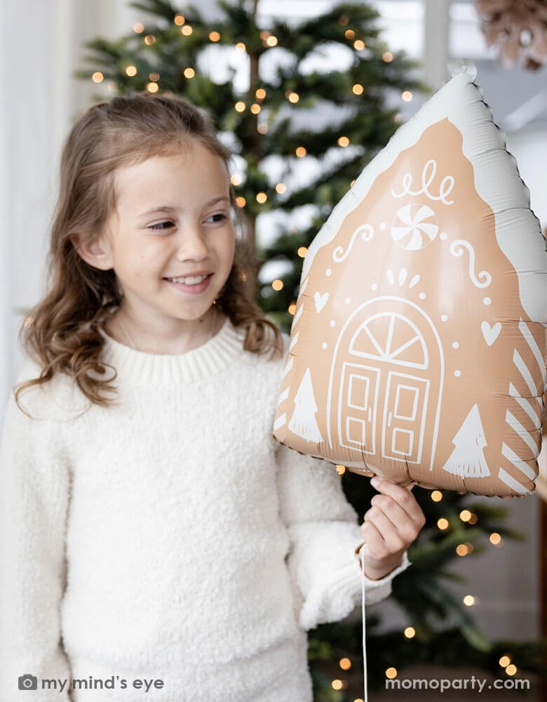 A girl in her cream white sweater holding Momo Party's 13.5" x 20" gingerbread house shaped foil balloon standing next to a big window. Behind her is a big Christmas tree lit up with small twinkling lights,