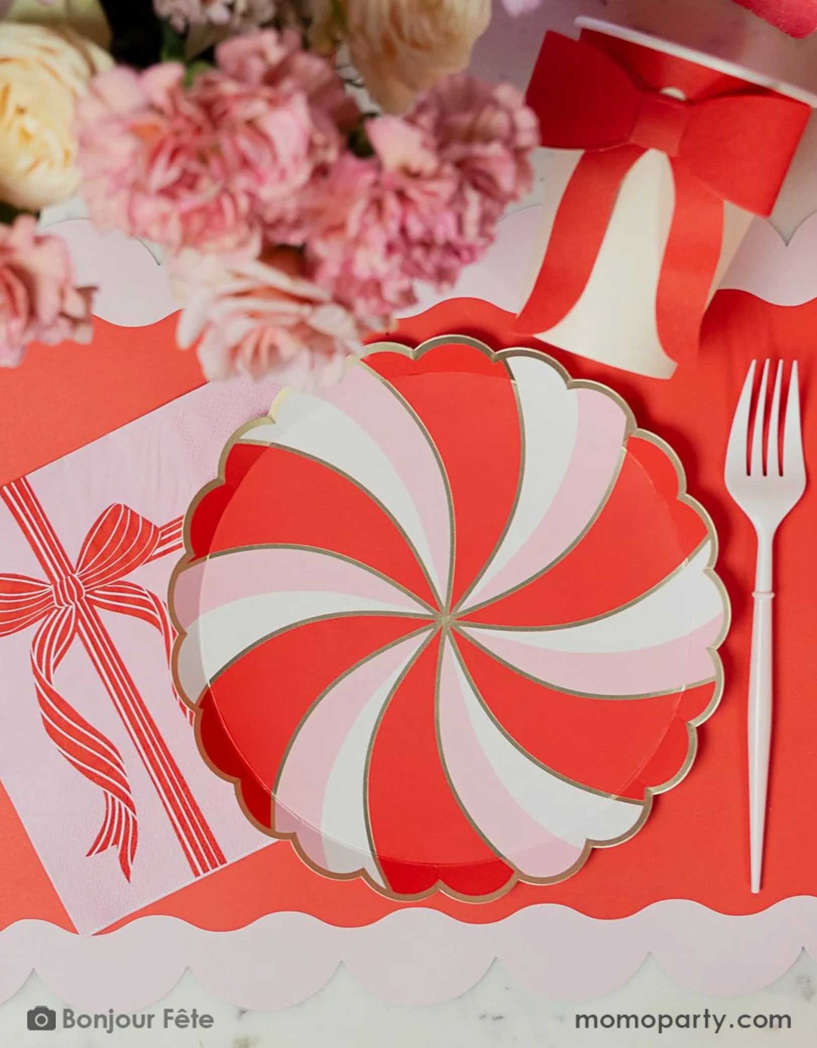 A festive Holiday party table setting featuring Momo Party's peppermint side plate with red and white candy swirl design, along with a pink large napkin with a red bow design and a pink party cup with red ribbon on a red scalloped shaped placemat with a floral arrangement around.