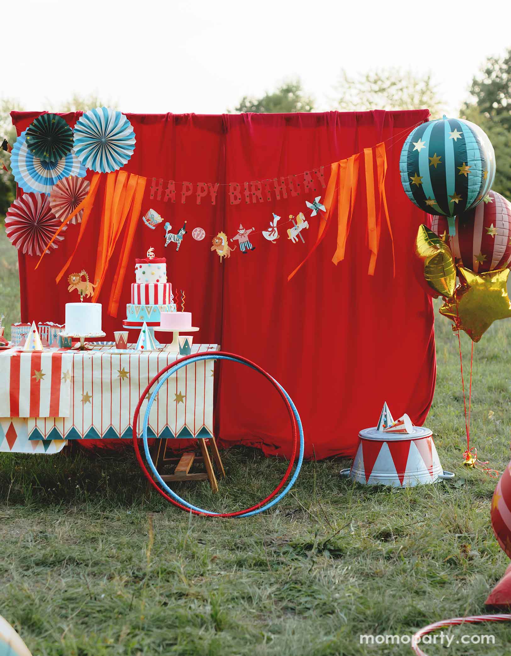 A vintage circus themed party set up in the park featuring various carnival circus themed party decorations from Momo Party including the red and green striped globe foil balloons along with gold star foil balloons, a red stage curtain decorated with Happy Birthday circus banner and colorful paper fans and a table filled with party tableware and multiple birthday cakes. Making this a whimsical and playful setting for a fun kid's circus birthday party.