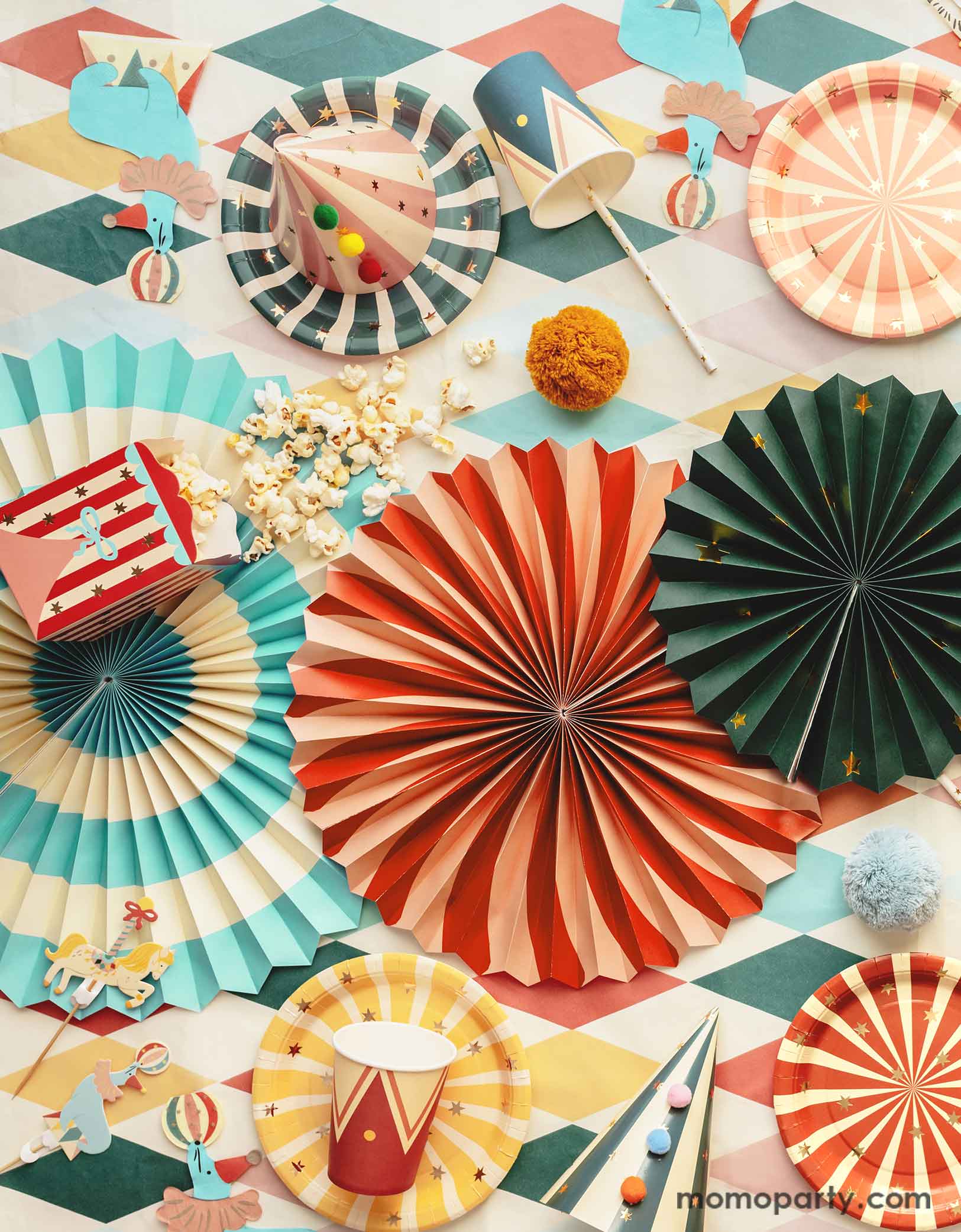 A carnival/circus themed part table featuring Momo Party's circus themed party supplies including colorful striped paper plates in 6 different colors, sea lion playing a ball shaped napkins, matching party cups, treat boxes, striped party hats with pom poms, circus themed cake toppers, and festive paper fans, with gold foil straws and popcorns on a circus themed striped table cover - making this great table setting idea for a fun and festive kid's carnival themed birthday celebration!