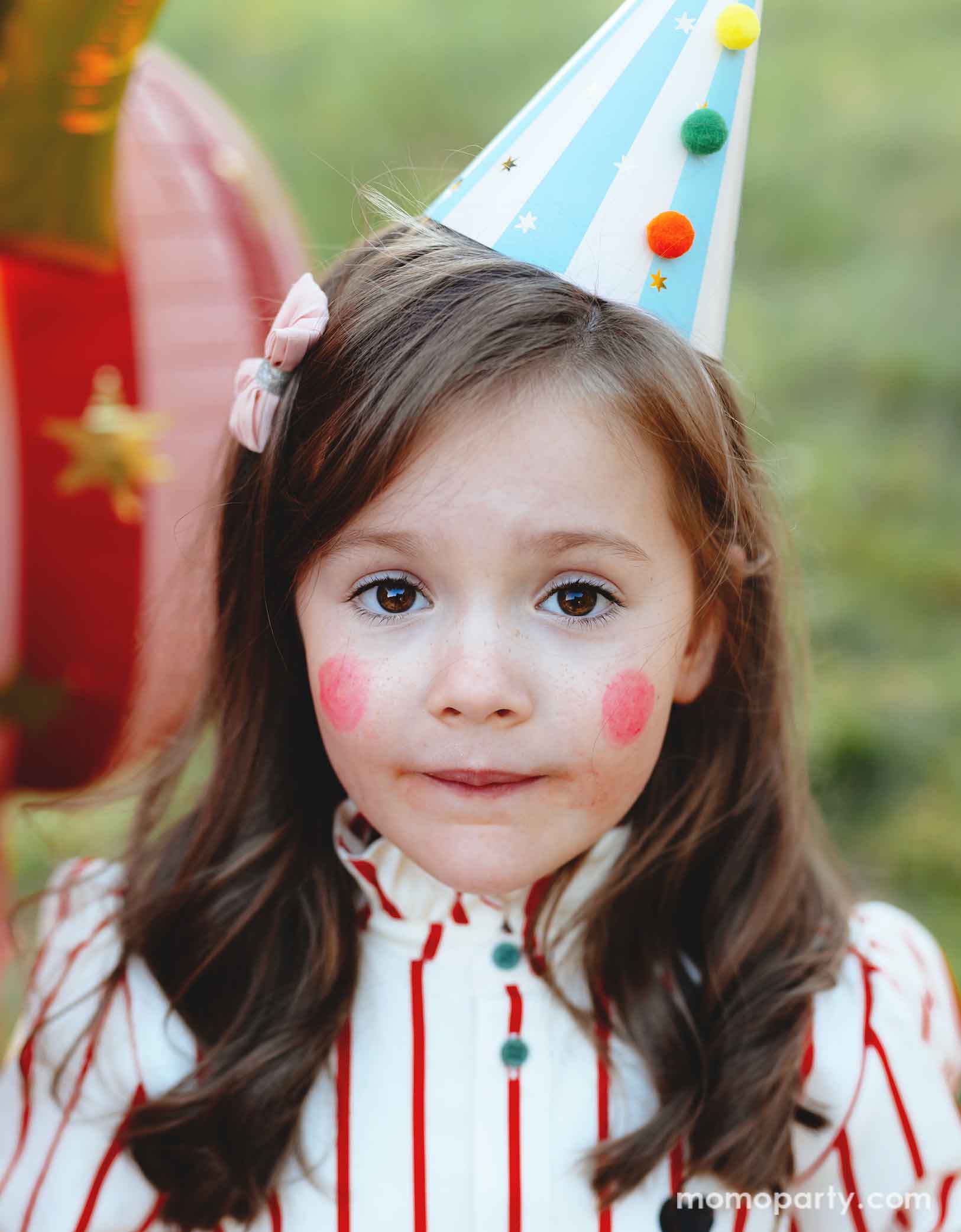 A girl dressed in vintage circus inspired outfits and make up wearing Momo Party's light blue striped Birthday Party Hat with Pompoms, in a festive carnival themed event. In her back there are some fun circus themed foil party balloons floating.