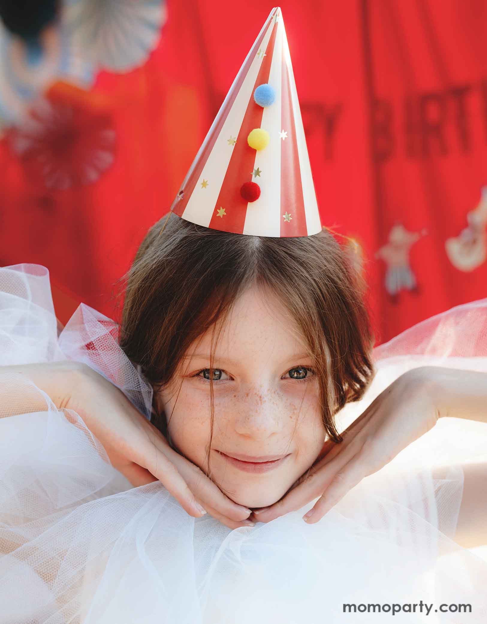 A girl dressed in dreamy white tulle dress wearing Momo Party's red striped Birthday Party Hat with Pompoms, in a festive carnival themed event. In her back is a red stage curtain decorated with circus themed birthday banner and colorful party fans.