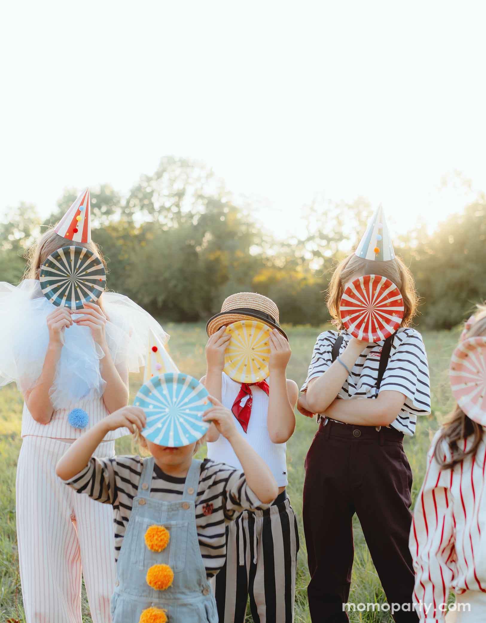 A bunch of kids dressed in the vintage circus outfits holding Momo Party's 7" Circus Striped Plates covering their faces. This set of 6 plates features vibrant stripes in 6 different colors and a charming gold star design, adding a touch of carnival magic to any meal. They're perfect additions to your circus or carnival themed party table!