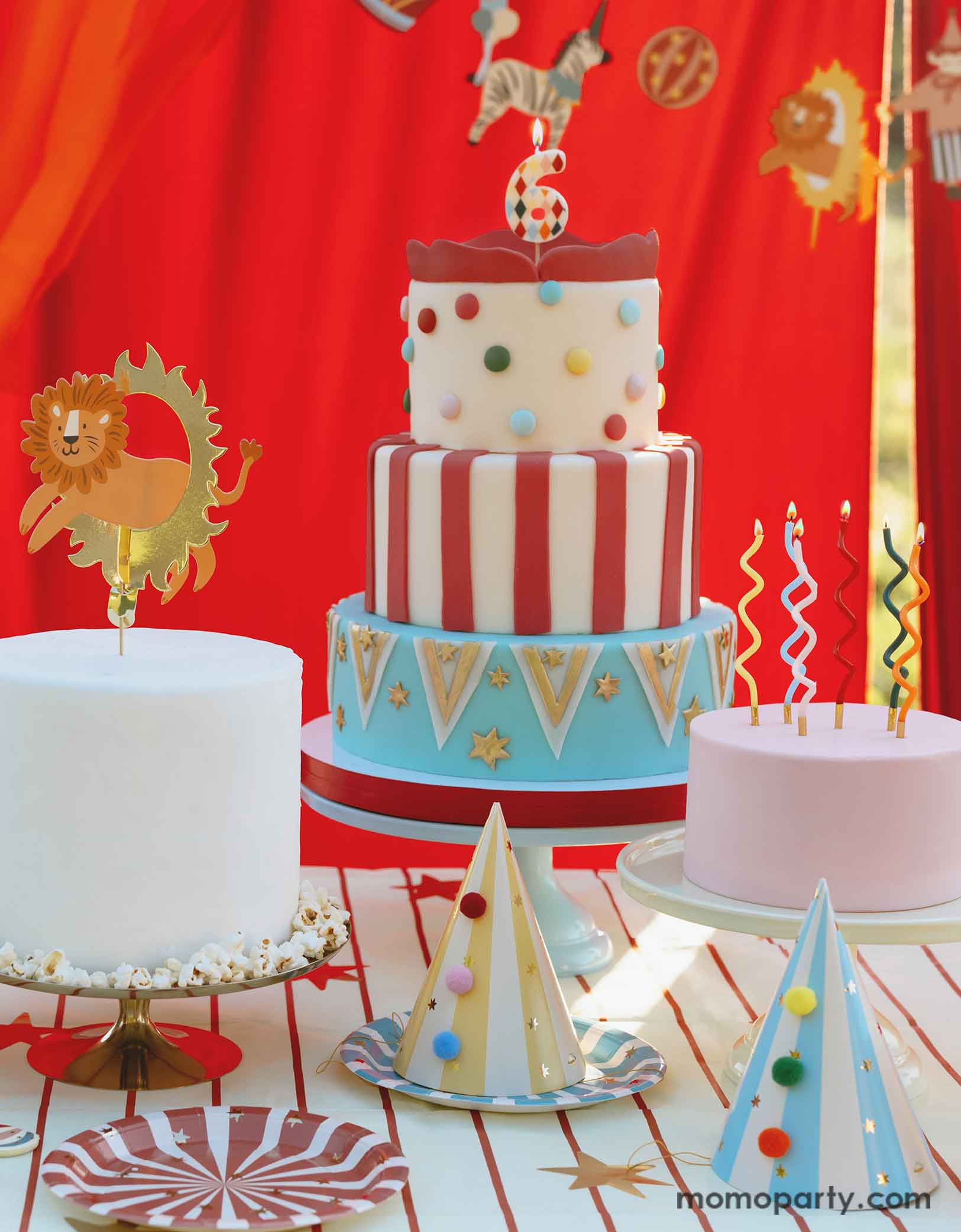 A circus party table featuring multiple carnival themed birthday cakes topped with fun candles and a lion jumping the loop cake topper. On the table there are Momo Party's circus themed party tableware including colorful striped paper plates in 6 different colors and striped party hats with pom poms. In the back there's a red curtain backdrop adorned with circus happy birthday banner, making this great table setting idea for a fun and festive kid's carnival/circus themed birthday celebration.