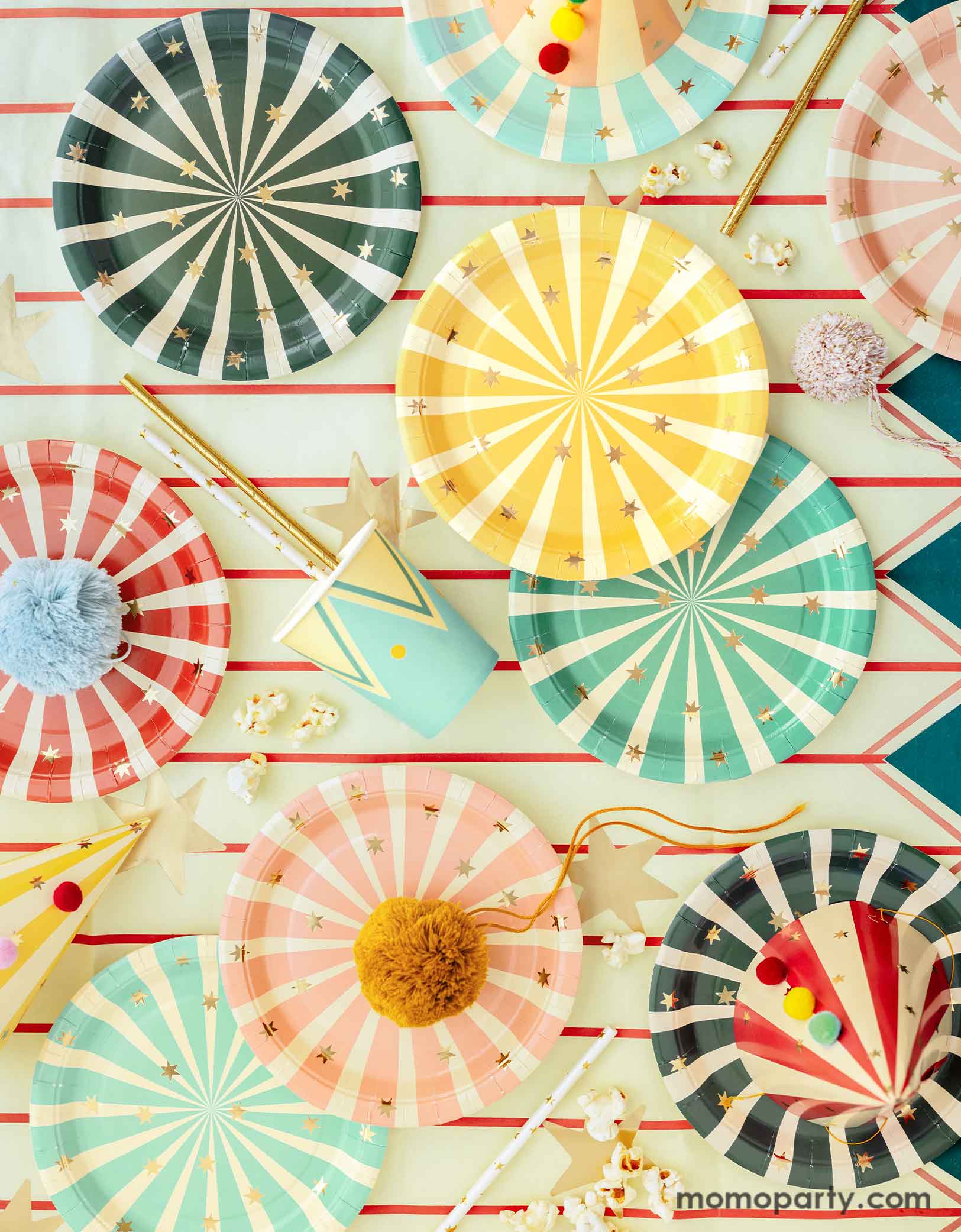 A carnival/circus themed part table featuring Momo Party's circus themed party tableware including colorful striped paper plates in 6 different colors, matching party cups, and striped party hats with pom poms, with gold foil straws and popcorns on a circus themed striped table cover - making this great table setting idea for a fun and festive kid's carnival themed birthday celebration!