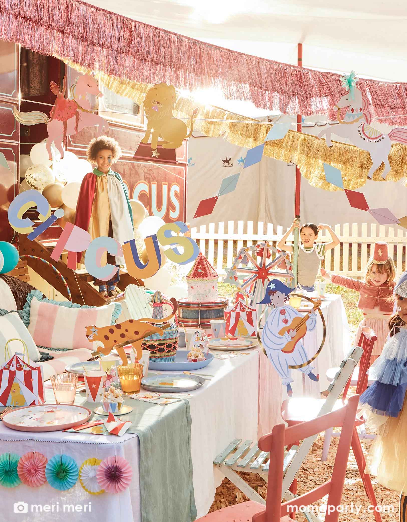 A festive circus themed party featuring Momo Party's circus themed party supplies including a table filled with circus themed party tablewares including plates, napkins, party cups, party bags and ferry's wheel cupcake stand. There's the circus party garland featuring circus animals with honeycomb decorations hung above the party table. Above the party garland, there are pink and gold fringe garlands. Around the table there are some kids dressed up in circus inspired costumes, making this a playful scene.