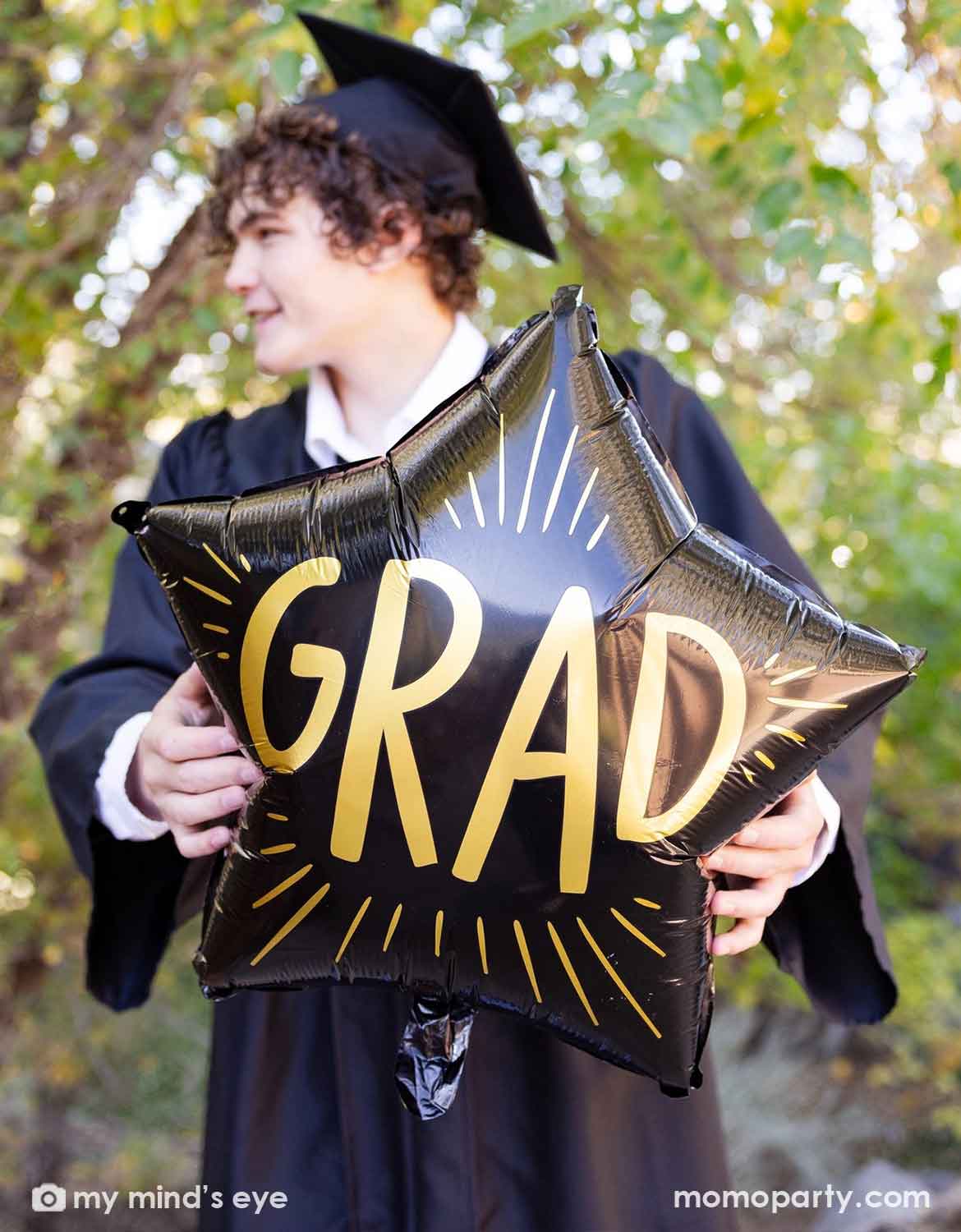 A collage grad in his graduation gown and cap holding Momo Party's 24" black star shaped foil balloon with 'GRAD' text on it with greenery in the background
