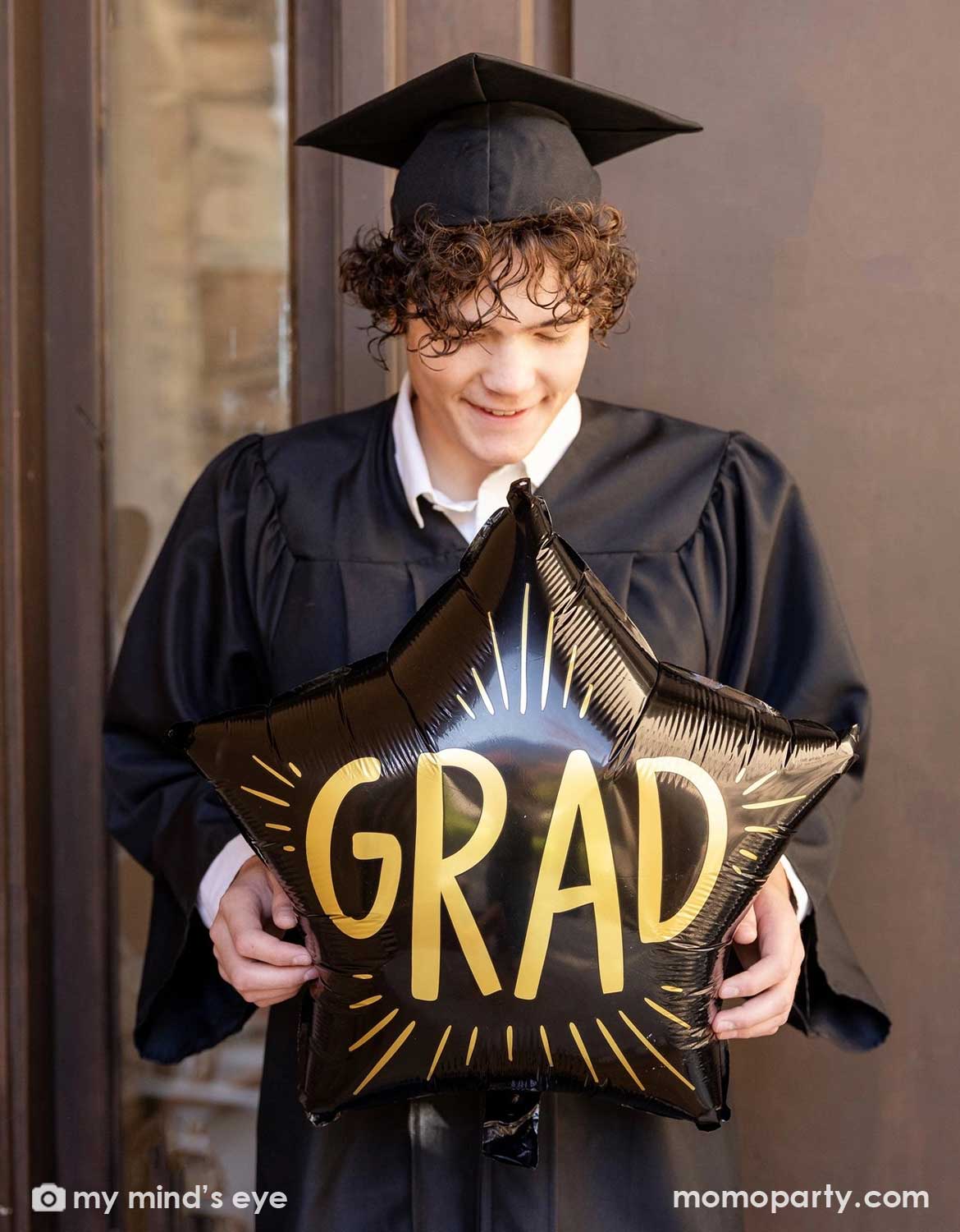 A collage grad in his graduation gown and cap holding Momo Party's 24" black star shaped foil balloon with 'GRAD' text on it standing by a black door at school.