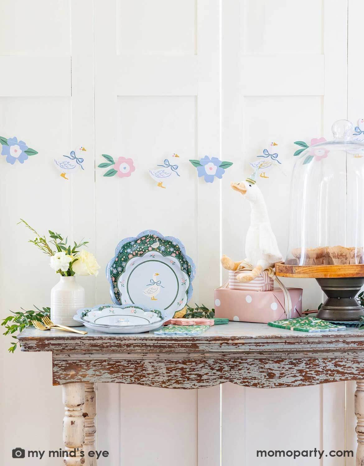 A rustic vintage style table setting featuring various goose themed party supplies and decorations from Momo Party including goose plates, napkins and party banner with floral design on a white wall background. On the table there are also some goose decoration and flowers, making this a perfect setting for kid's Silly Goose themed birthday party, a baby shower or an Easter celebration.