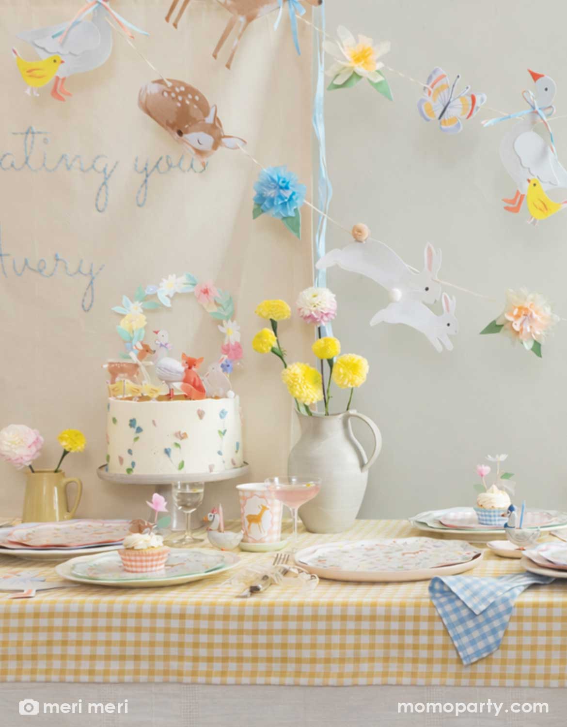 A rustic cottage themed table setting with little woodland animals of deer, goose, bunny, fox, chicks in tablewares and decorations from Momo Party - including floral little animal cake toppers, party cups, matching plates and napkins and an adorable party garland hung above the table covered with a soft gingham tablecloth. With spring florals in soft yellow and pink on the table, this table is perfect for a baby shower, an Easter celebration or young kid's storybook rustic cottage inspired birthdays.