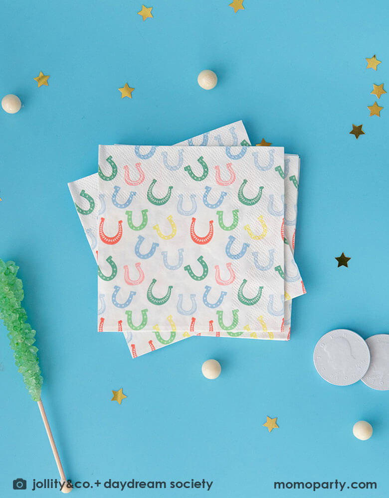 Momo Party's 5" x 5" horseshoe small napkins by Daydream Society on a bright blue table with gold star foil confetti and white gum balls surrounding them. With a green rocky ice lollipop and white chocolate coins next to the napkins.