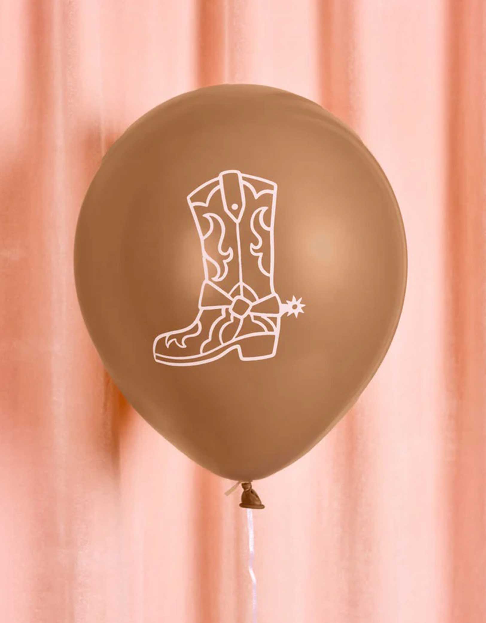 A 12" brown latex balloon with a white cowboy boot design from Momo Party on a pink curtain background.