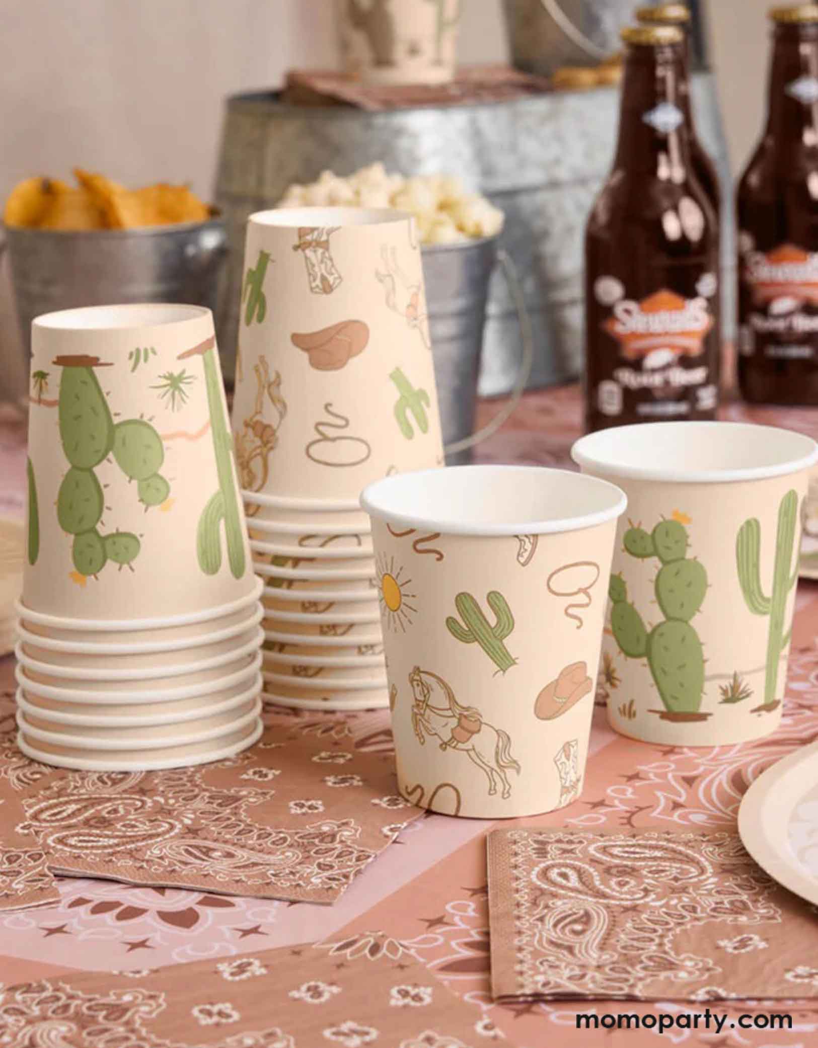 Stacks of Momo Party's 9oz Wild Wild West Party Cups with Western cowboy and desert, cactus design sitting on a set of brown small napkins with bandana pattern with beer bottles and snacks in the metal buckets on a cowboy rodeo themed party table.