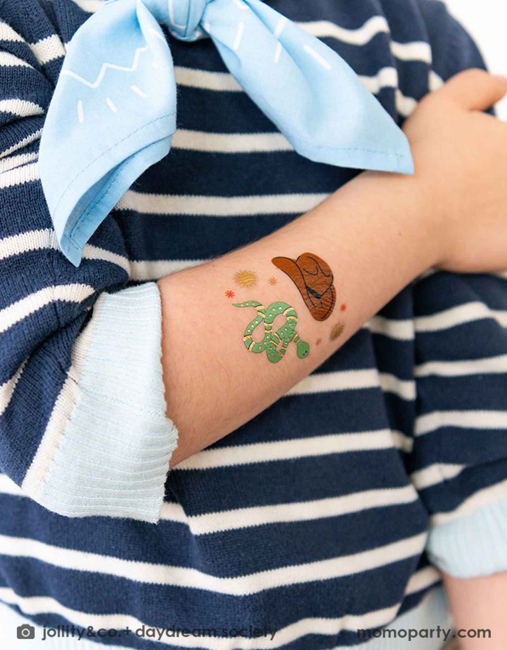 A boy wearing navy striped shirt with a light blue bandana. On his arm he has Momo Party's cowboy hat & snack temporary tattoos on it. This temporary tattoo set is a great party favor for kid's Western or cowboy themed birthday party.