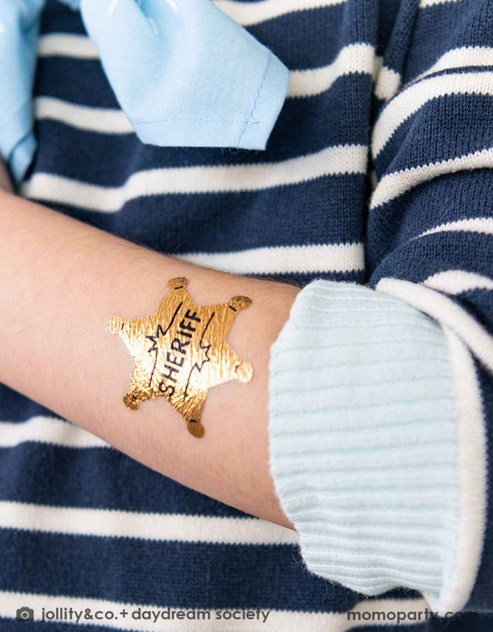 A boy wearing navy striped shirt with a light blue bandana. On his arm he has Momo Party's sheriff badge temporary tattoo on it. It's a great party favor for kid's Western or cowboy themed birthday party.