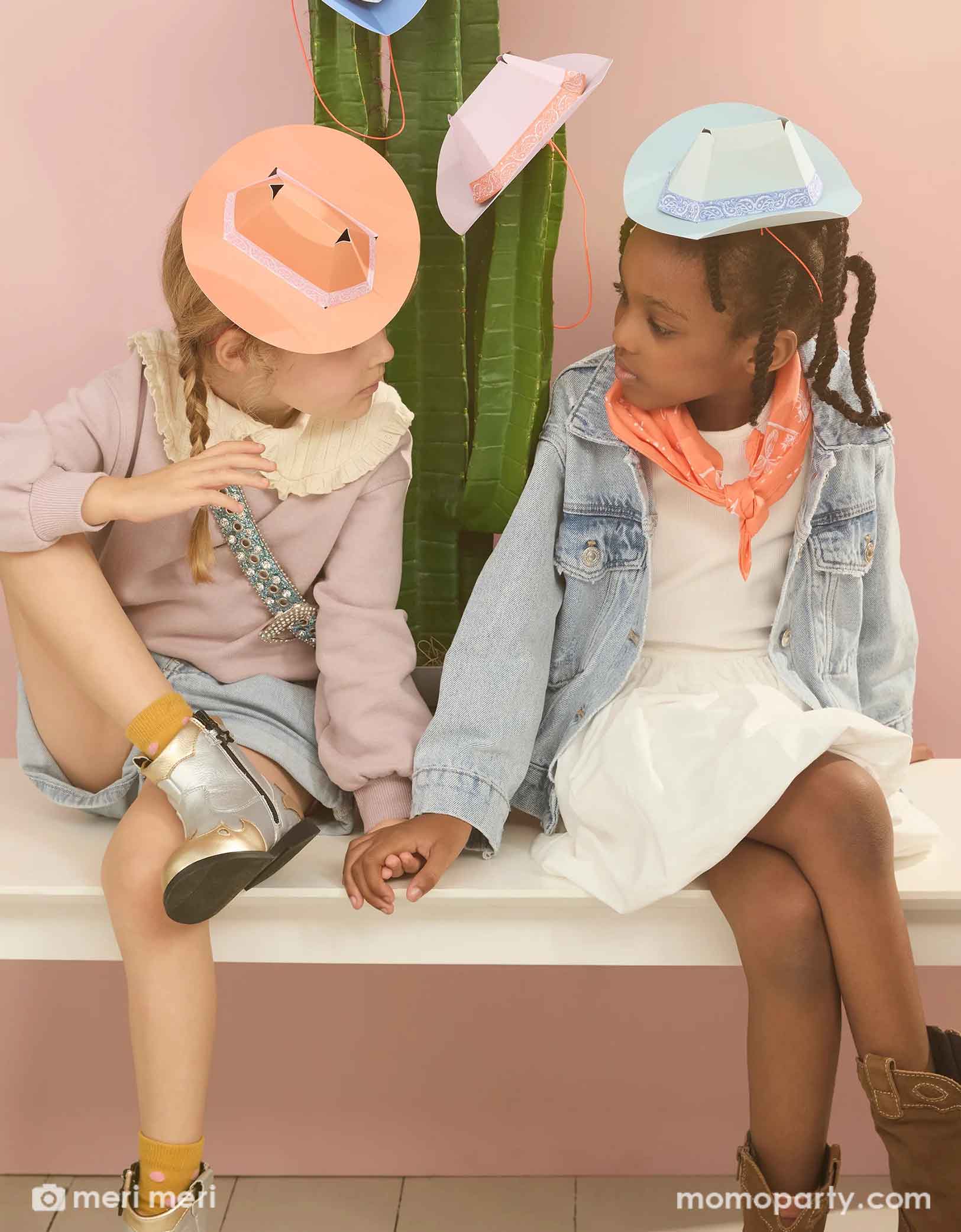 Two school aged girls dressed in Western inspired outfits including bandanas and  cowboy boots, sitting on a white bench in front of cactus plant. Each of them wear the Meri Meri's 6.3" mini Western party hat, one in light blue, one in orange. There are also a couple more hats hung on the cactus plant.