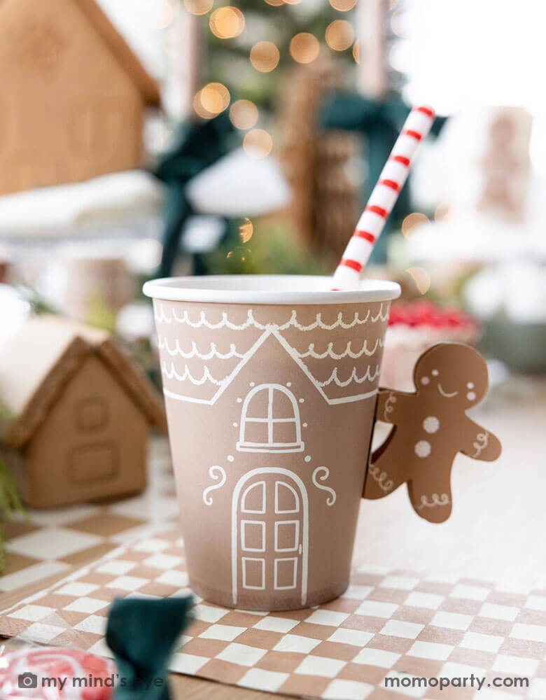 A festive gingerbread themed Holiday party table set up featuring gingerbread themed party tableware by Momo Party including the 12 oz Gingerbread Handled Paper Cup with a festive gingerbread man handle, the mocha checkered napkins and paper table runner. With gingerbread houses and Christmas trees in the back this creates a cozy yet festive vibe for a whimsical Holiday party for kids.